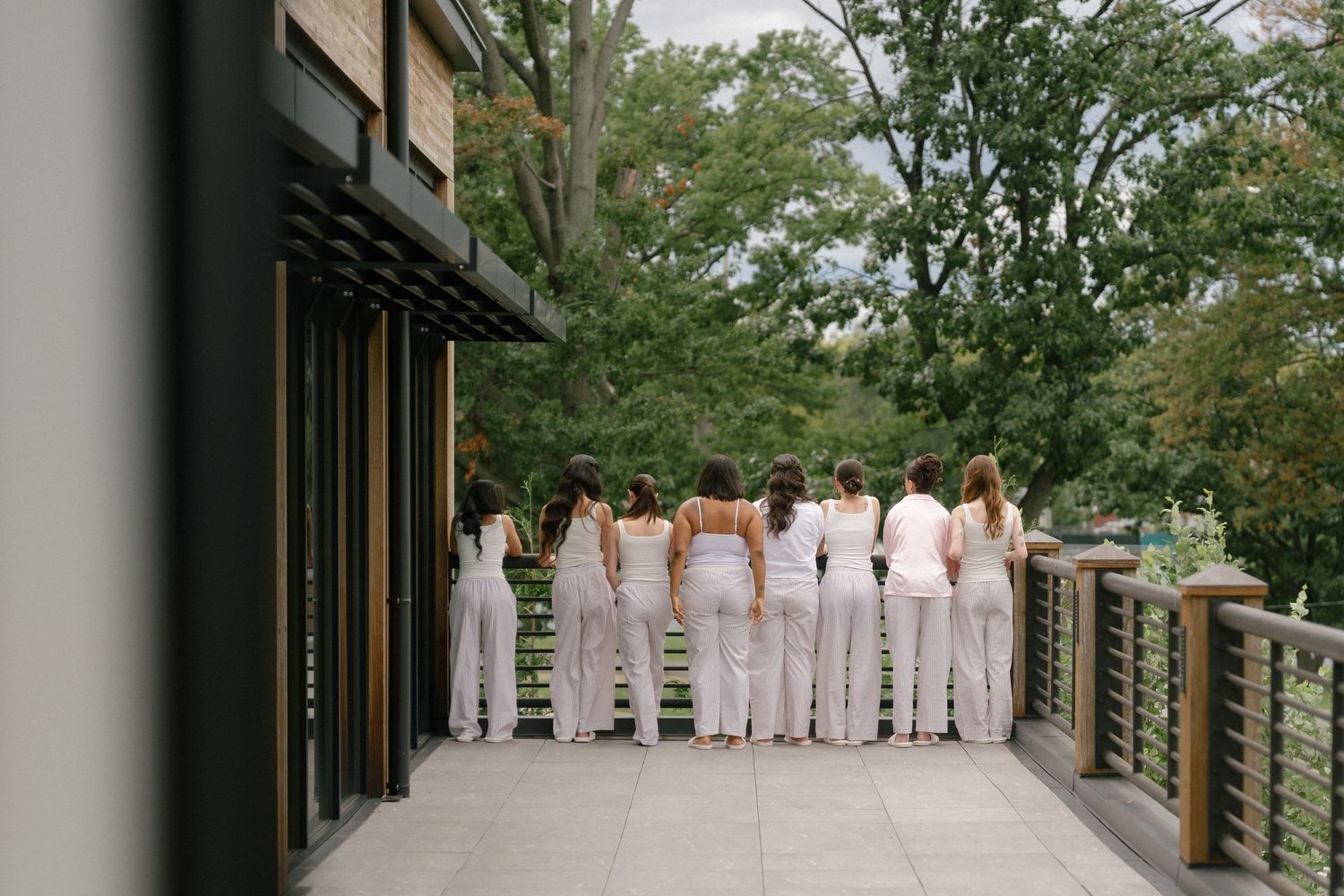 Bridesmaids in white outfits standing together on outdoor deck surrounded by lush greenery.