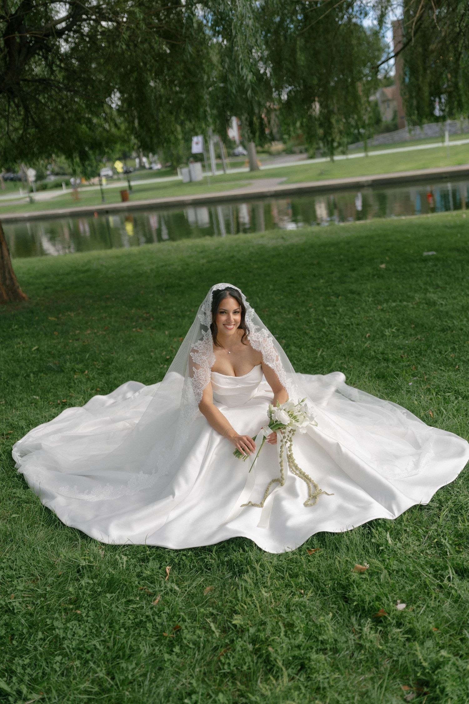 Radiant bride in strapless ball gown with lace veil sitting on green grass holding white floral bouquet outdoors.
