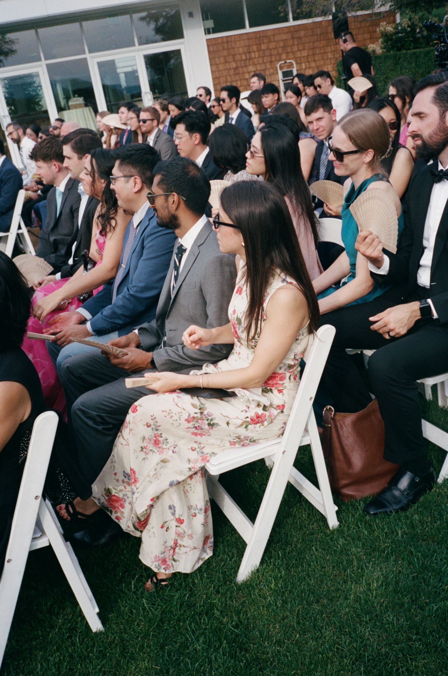 Wedding guests seated in white chairs at outdoor ceremony, dressed in formal and semi-formal attire.
