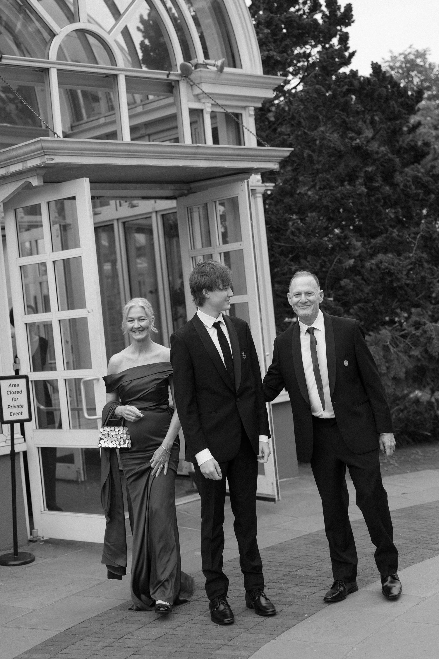 Elegant family of three poses in formal attire outside a glass conservatory building in black and white photo.