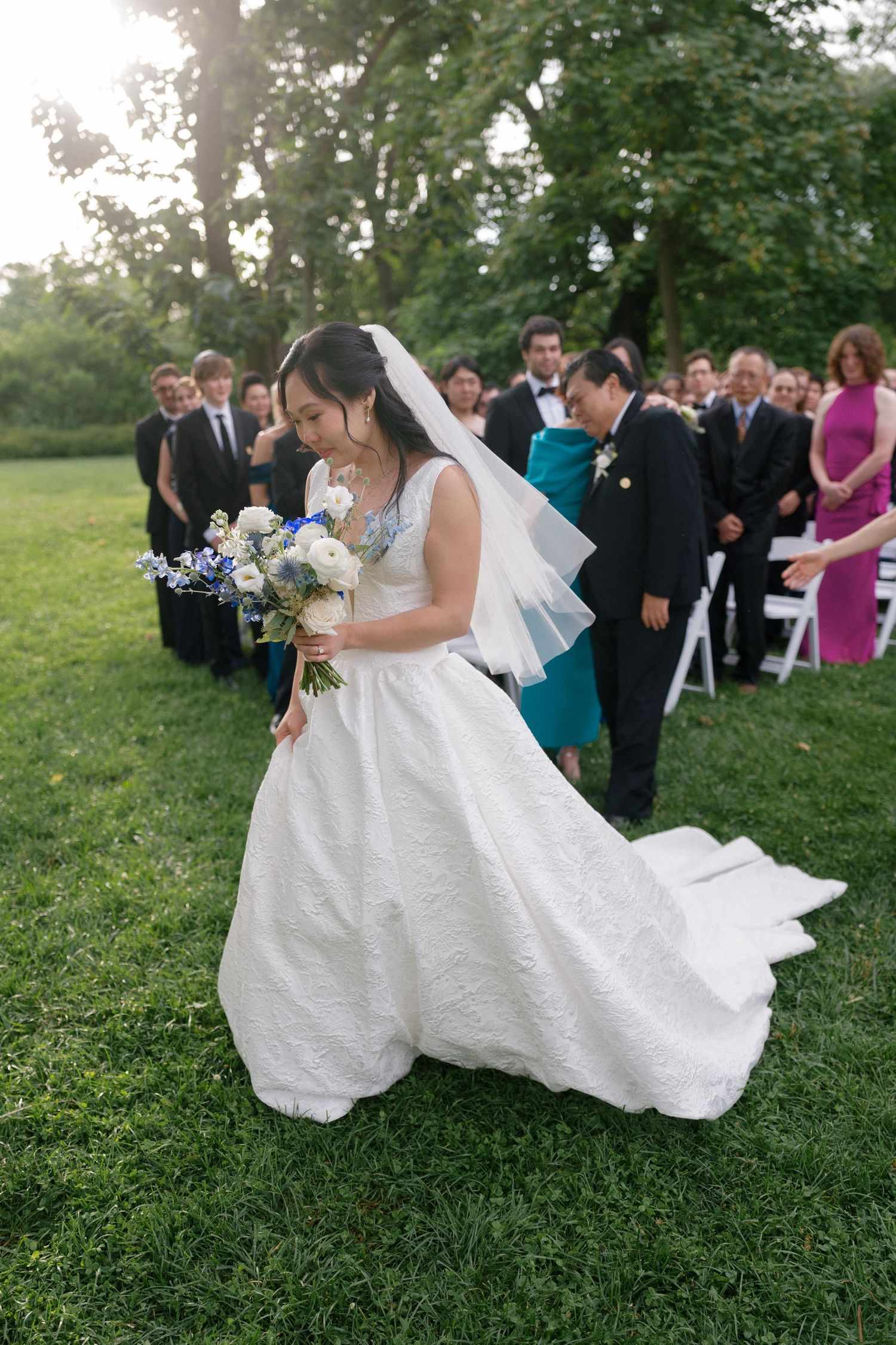 Bride in white gown holding bouquet walks down outdoor aisle.