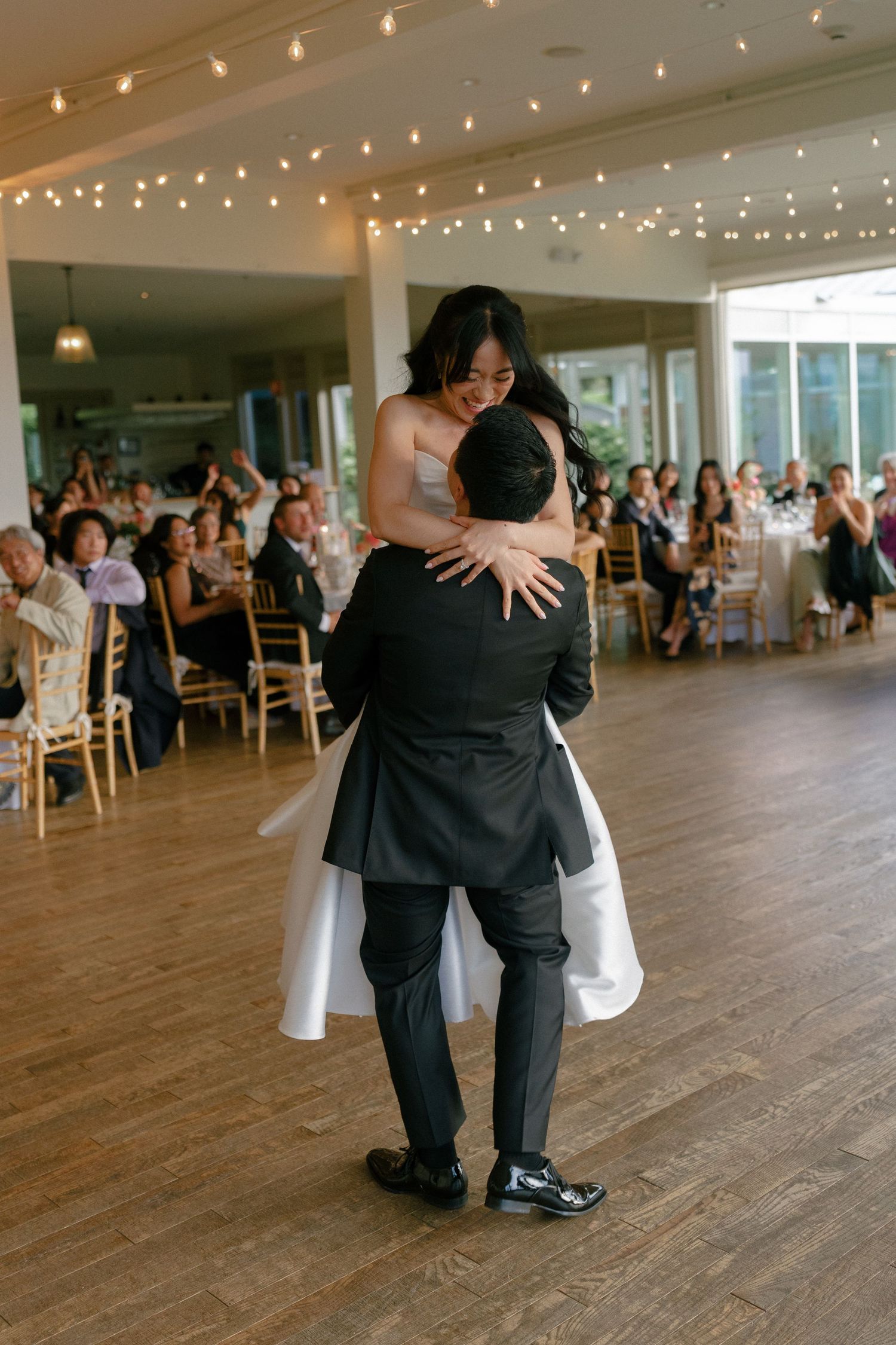Groom lifts bride in white gown during first dance in elegant reception hall with string lights.