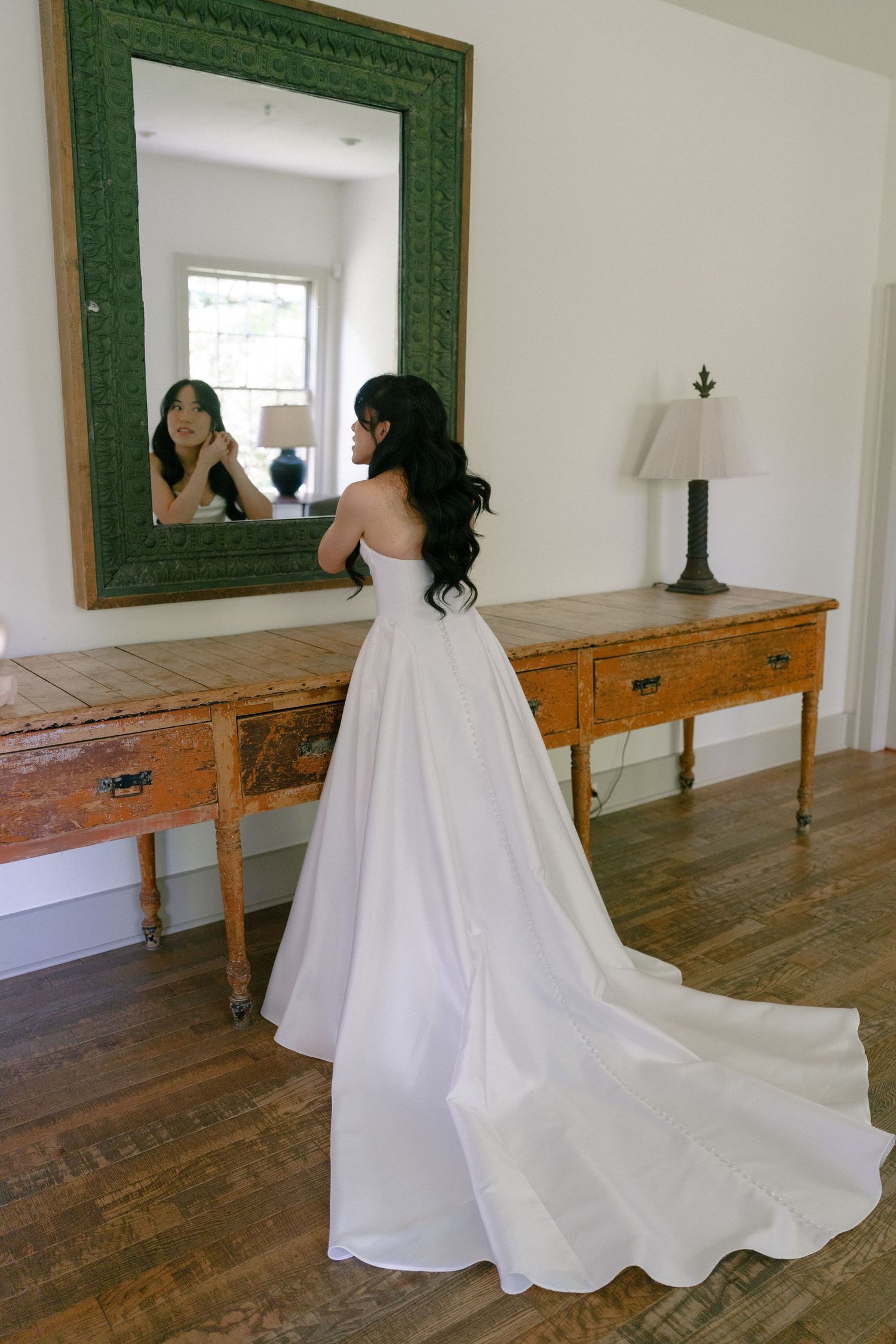Bride in strapless white ball gown with long train stands before ornate mirror in rustic room.