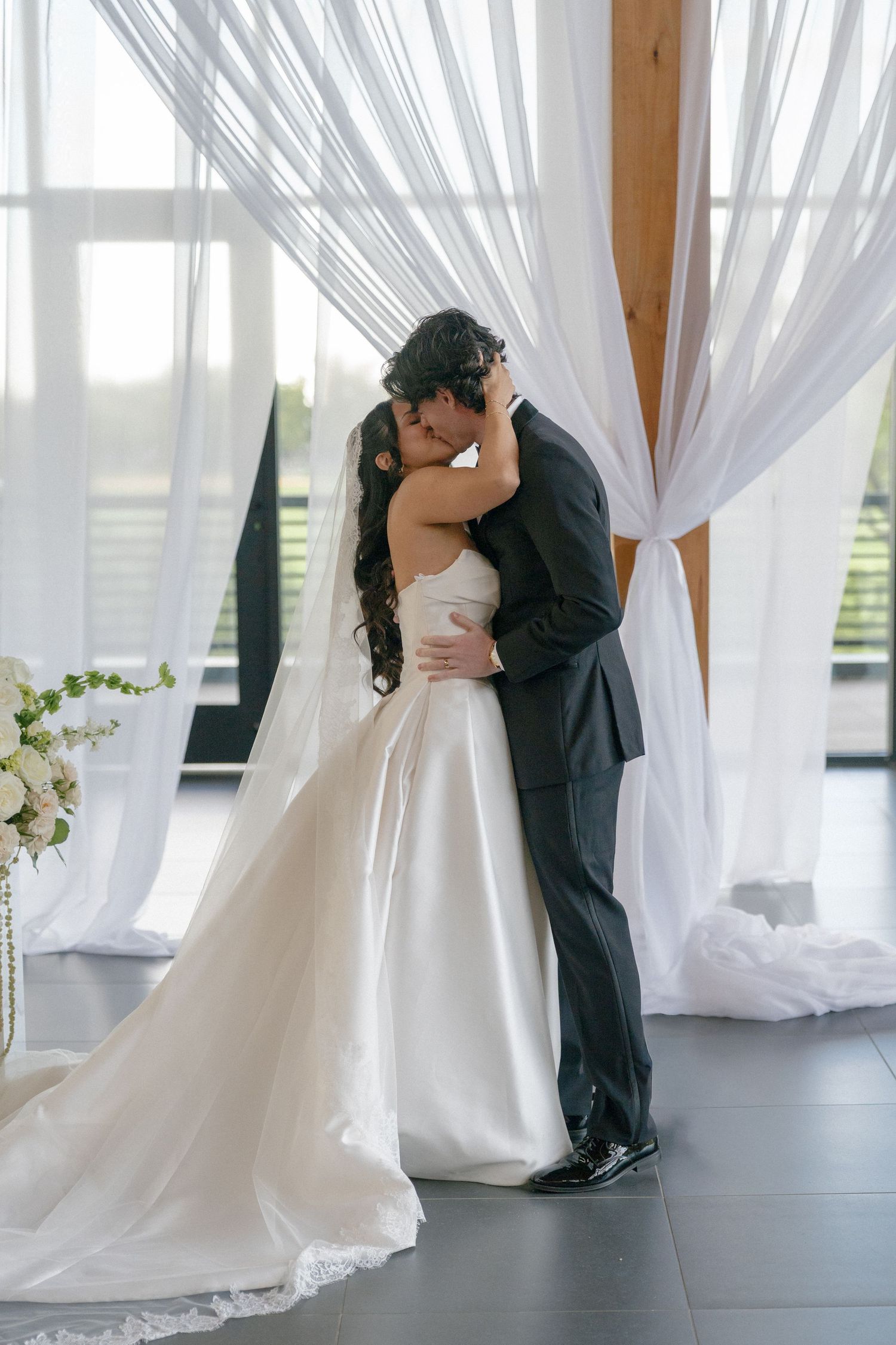 A bride and groom share their first kiss at the altar, framed by elegant white draping curtains in a bright wedding venue.
