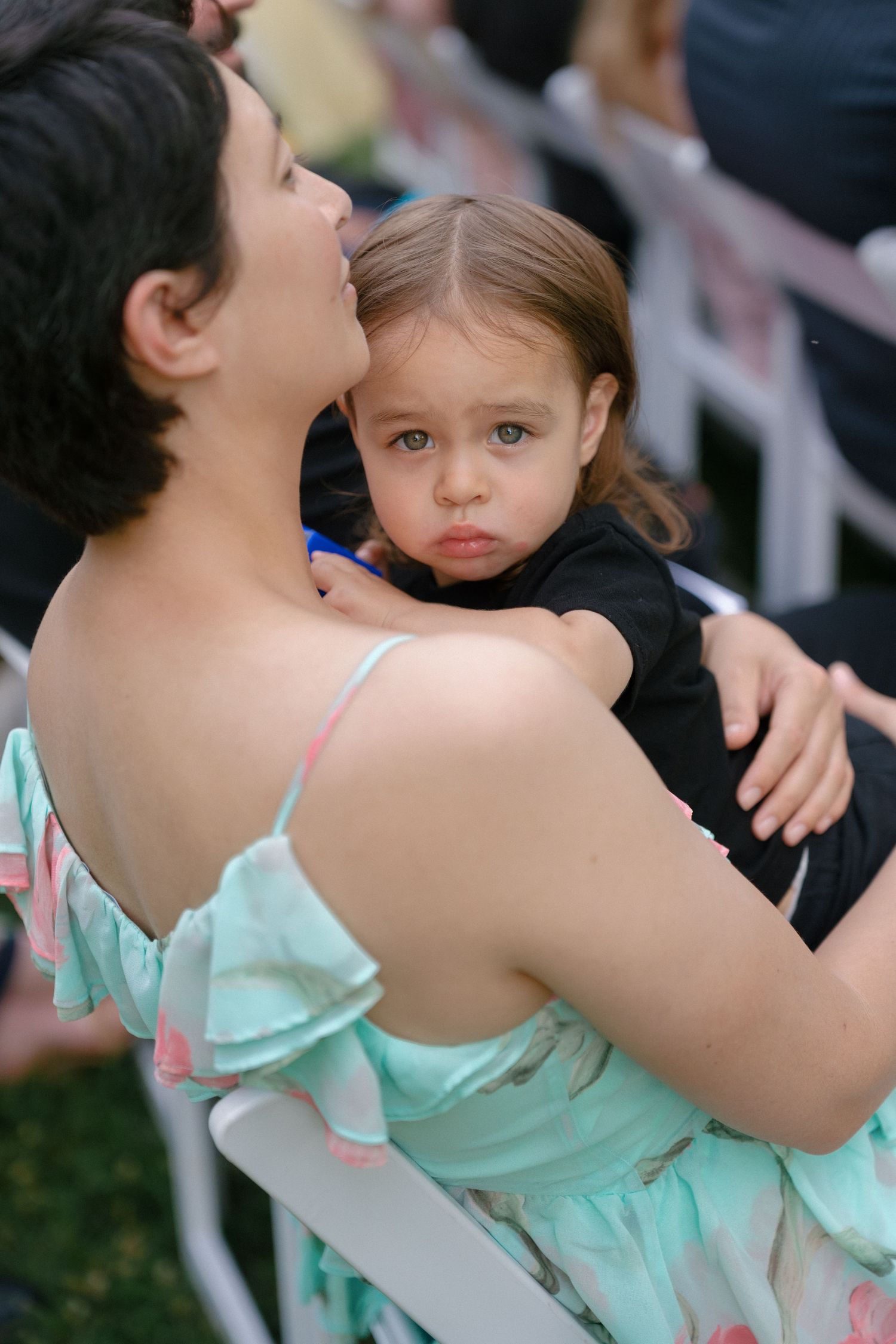 A mother in a teal floral dress holds her toddler daughter at an outdoor wedding ceremony.