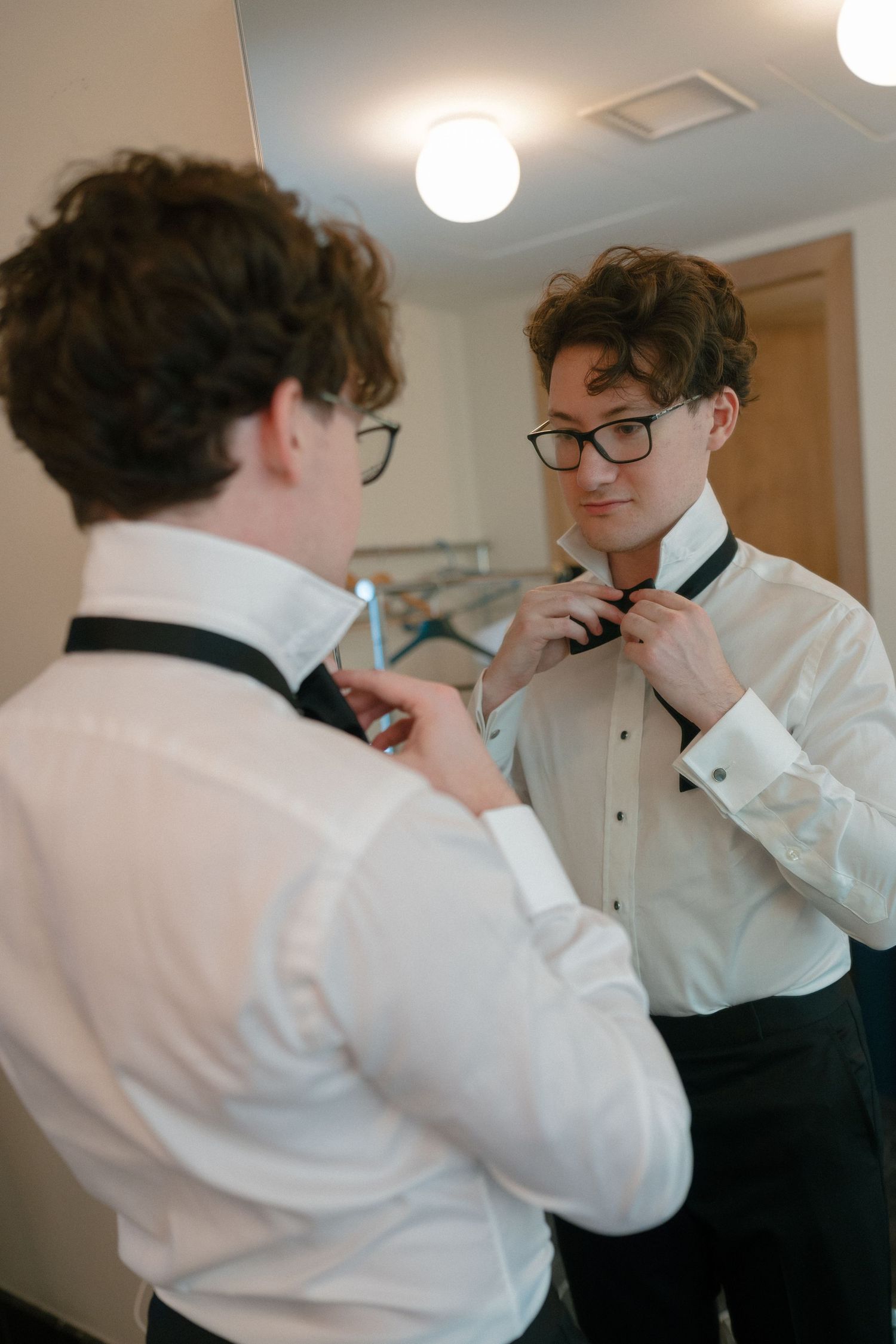 Groom in white dress shirt adjusts his bow tie while looking in a mirror during wedding day preparations.