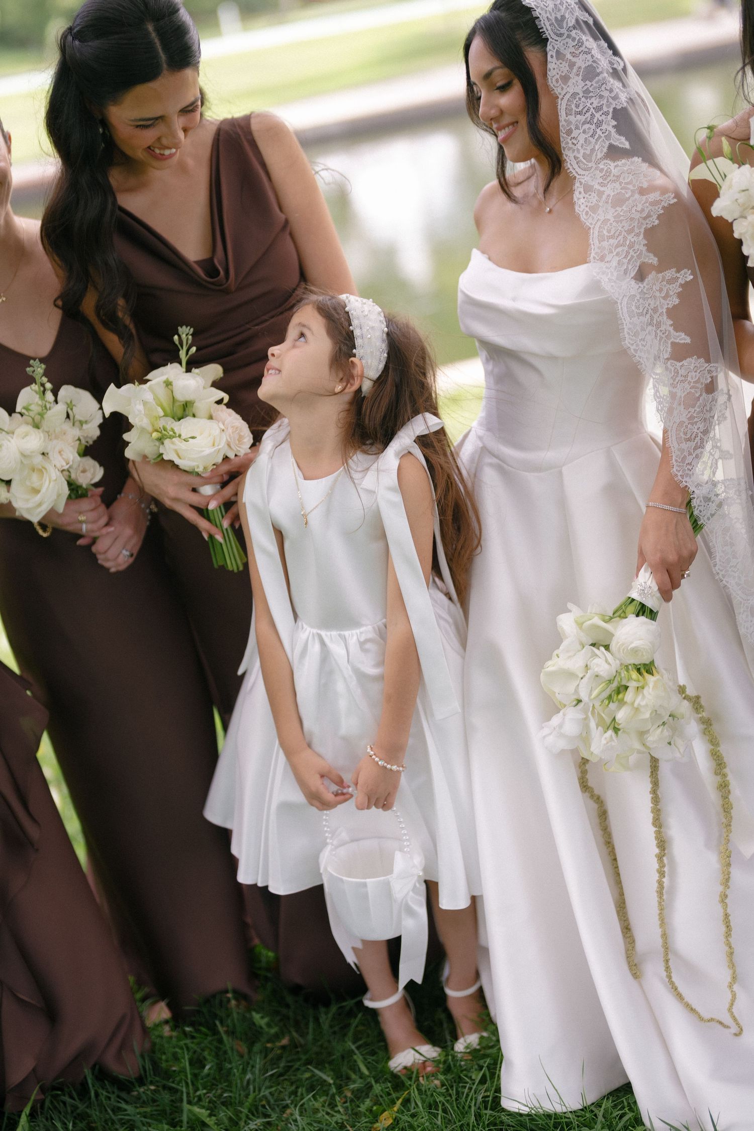 Bride in lace veil walks with flower girl and bridesmaids in brown dresses outdoors.