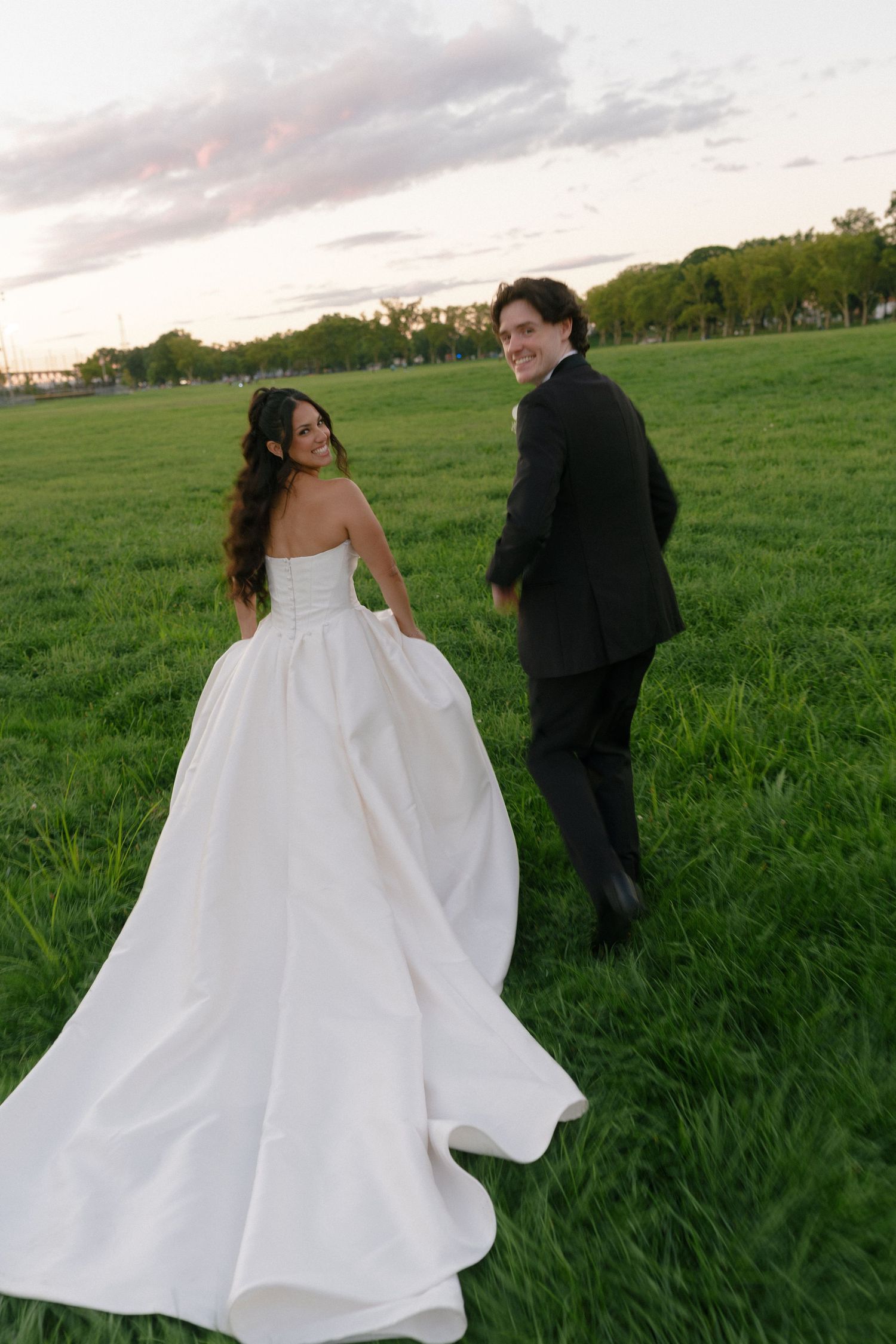 Bride in white ballgown and groom in black suit walk through green field at sunset, smiling back at camera.