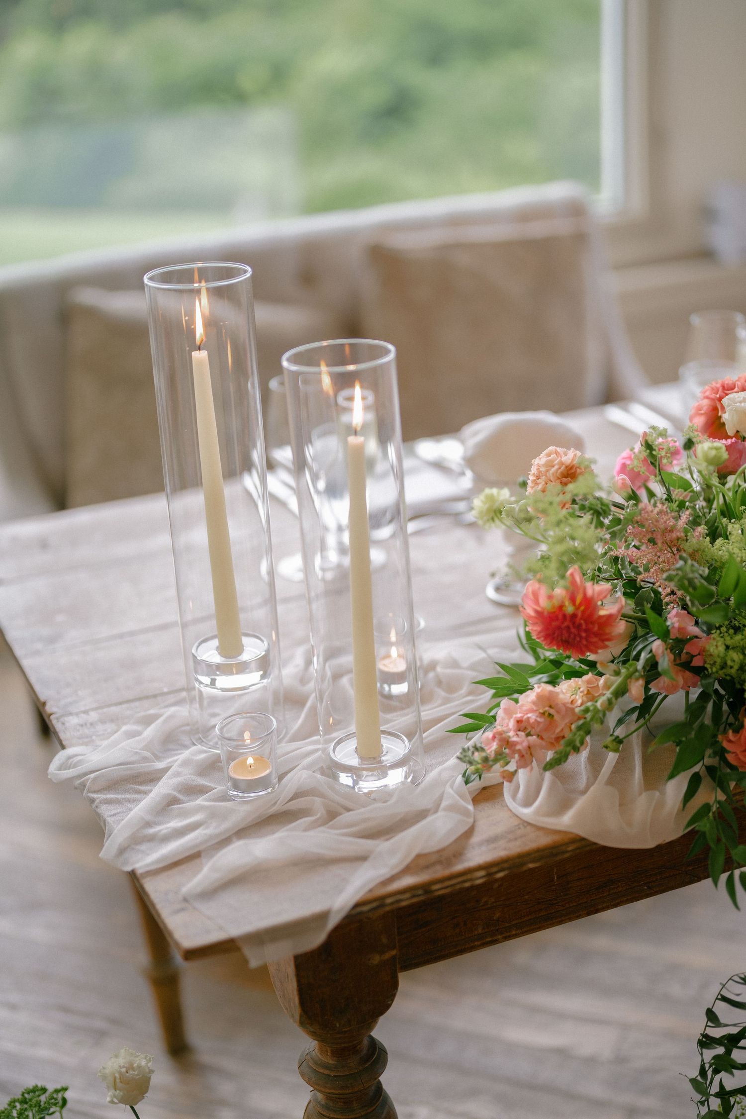 Elegant wedding table decor with tall glass candle holders and pink floral centerpiece near window.