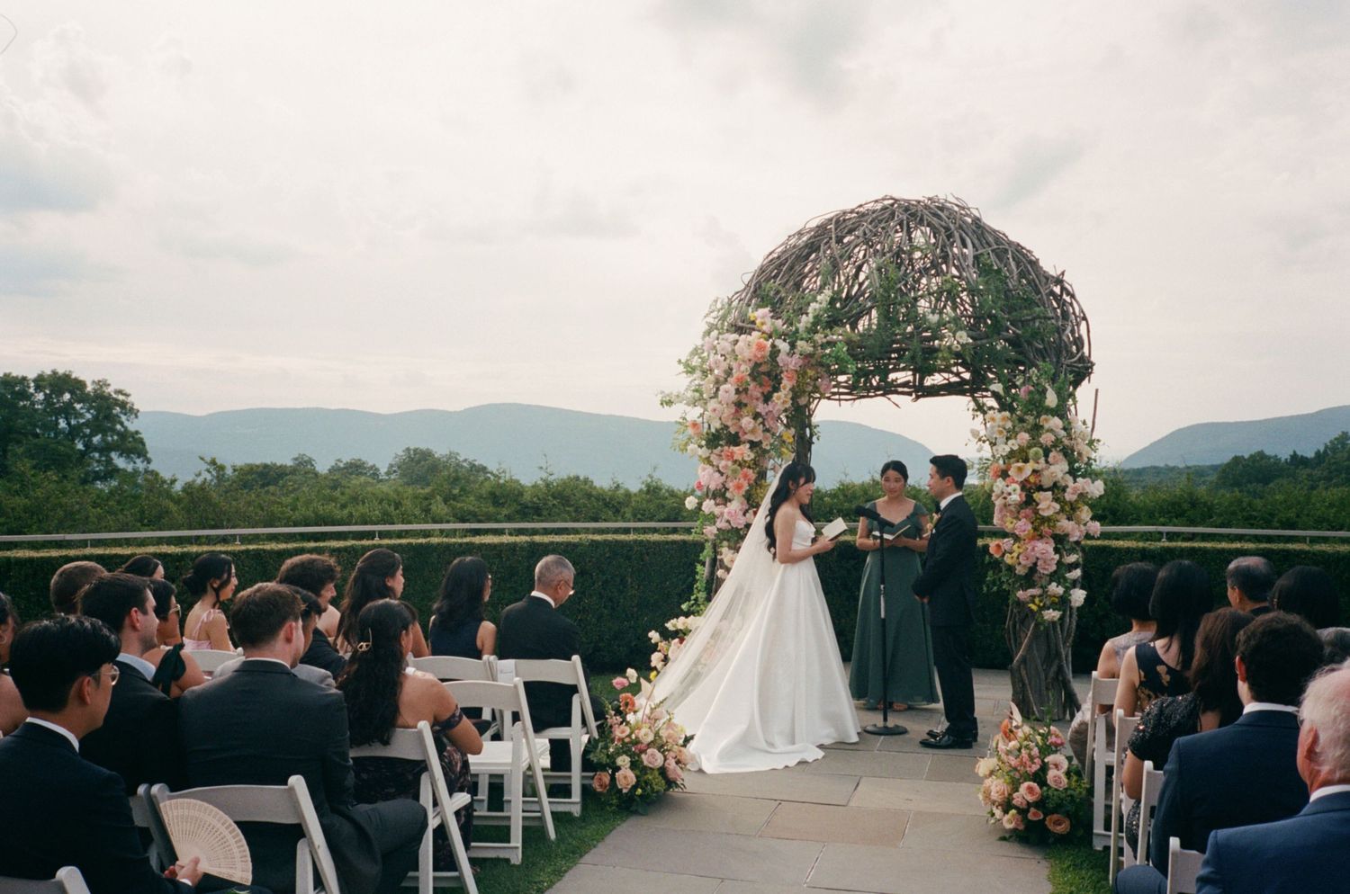 Outdoor wedding ceremony under floral arch with mountain views, guests seated on white chairs.