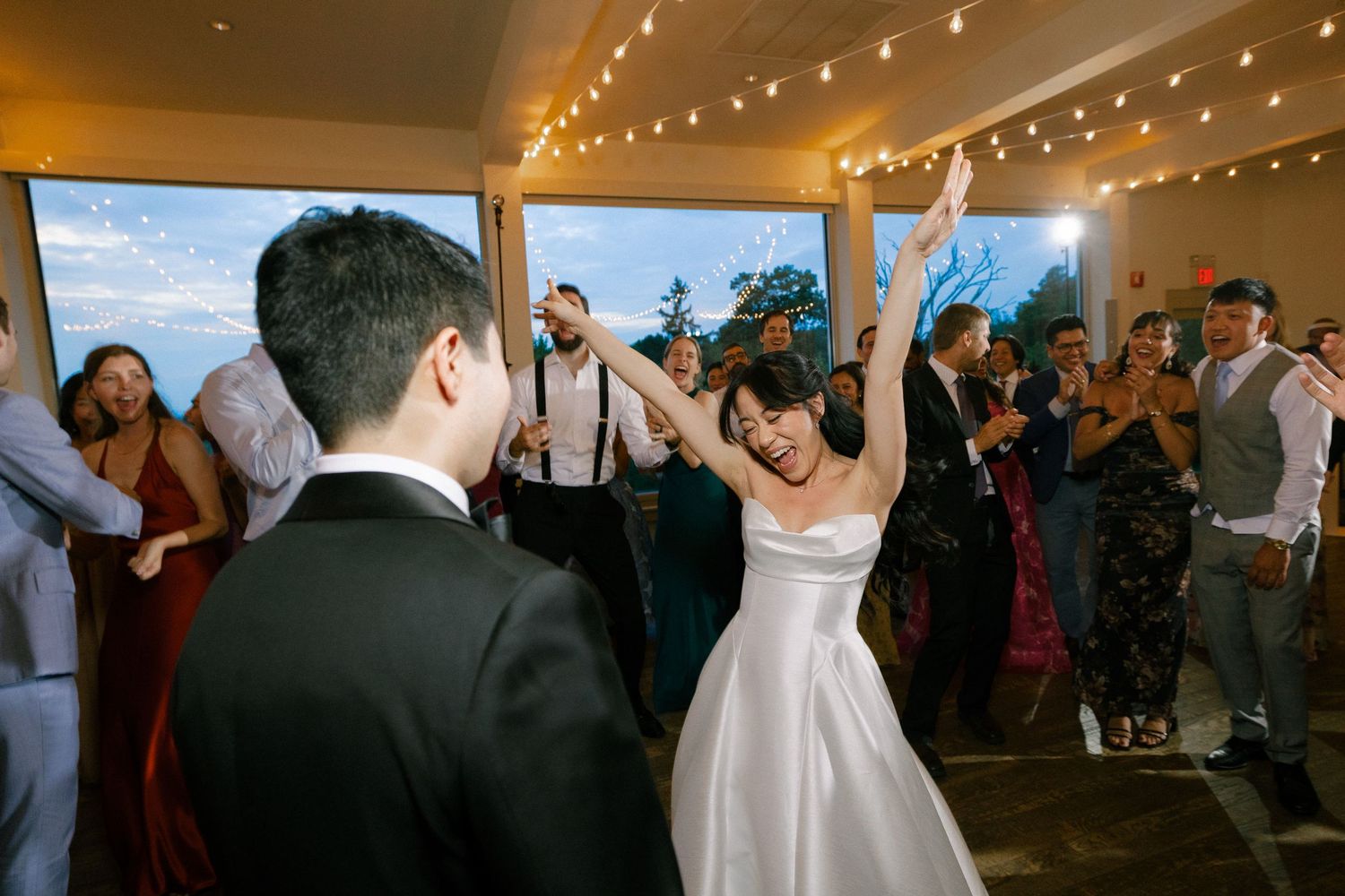 Bride and groom share first dance surrounded by cheering wedding guests in elegant venue with string lights.