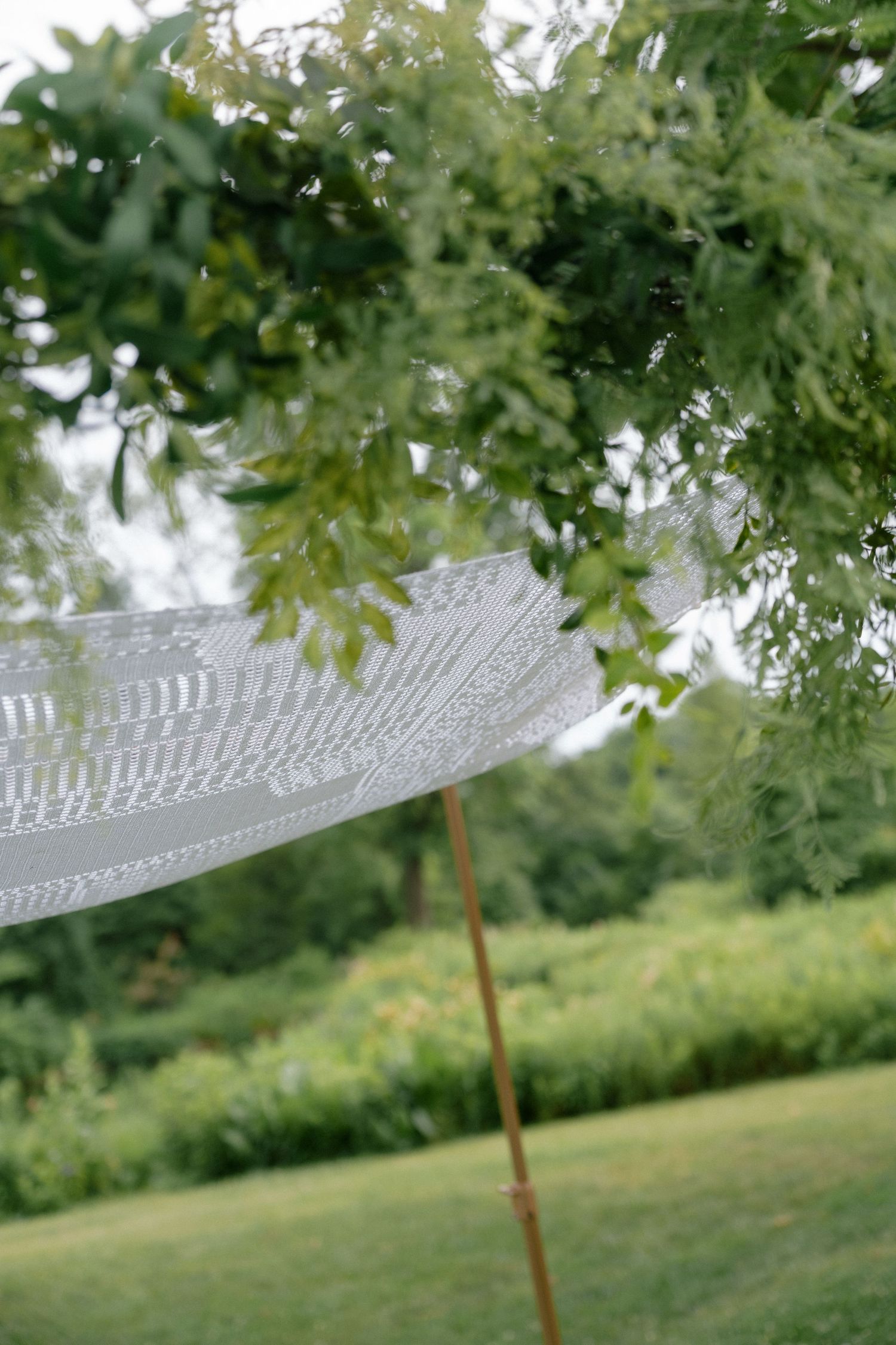 Delicate white lace wedding canopy on a wooden pole surrounded by green trees outdoors.