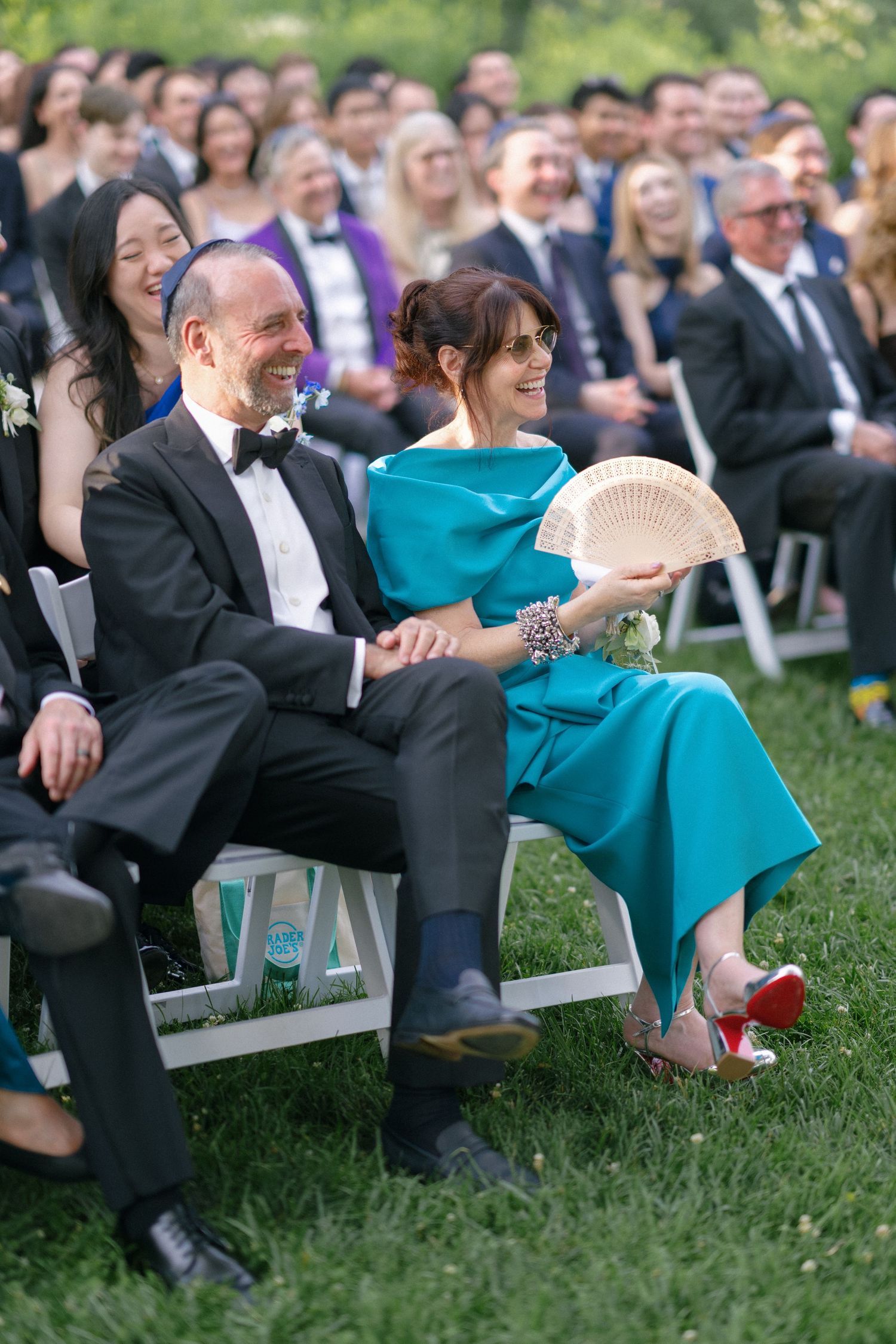 Wedding guests laugh and smile during outdoor ceremony, woman in teal dress holds decorative fan.