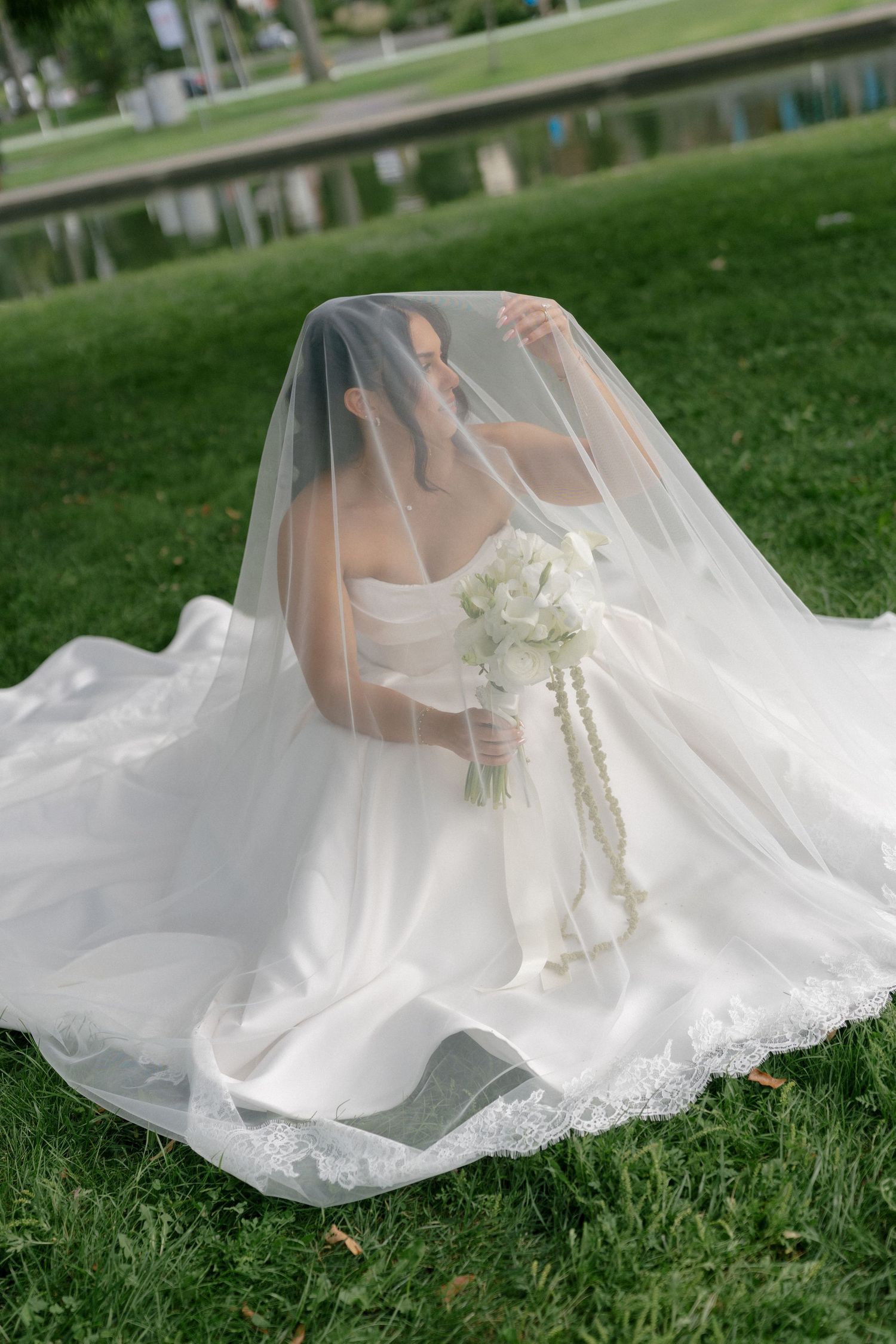 Bride sitting on grass with veil draped over her, holding a white floral bouquet.