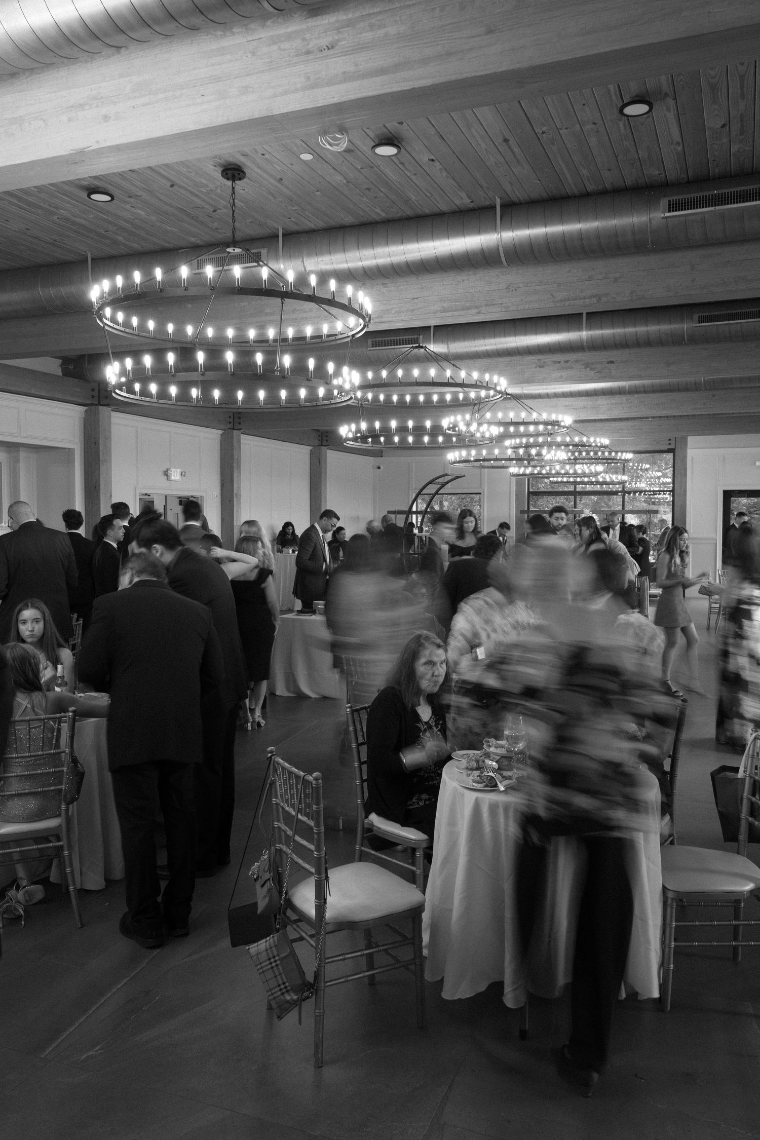 Black and white photo of wedding guests mingling under circular chandeliers in reception hall.