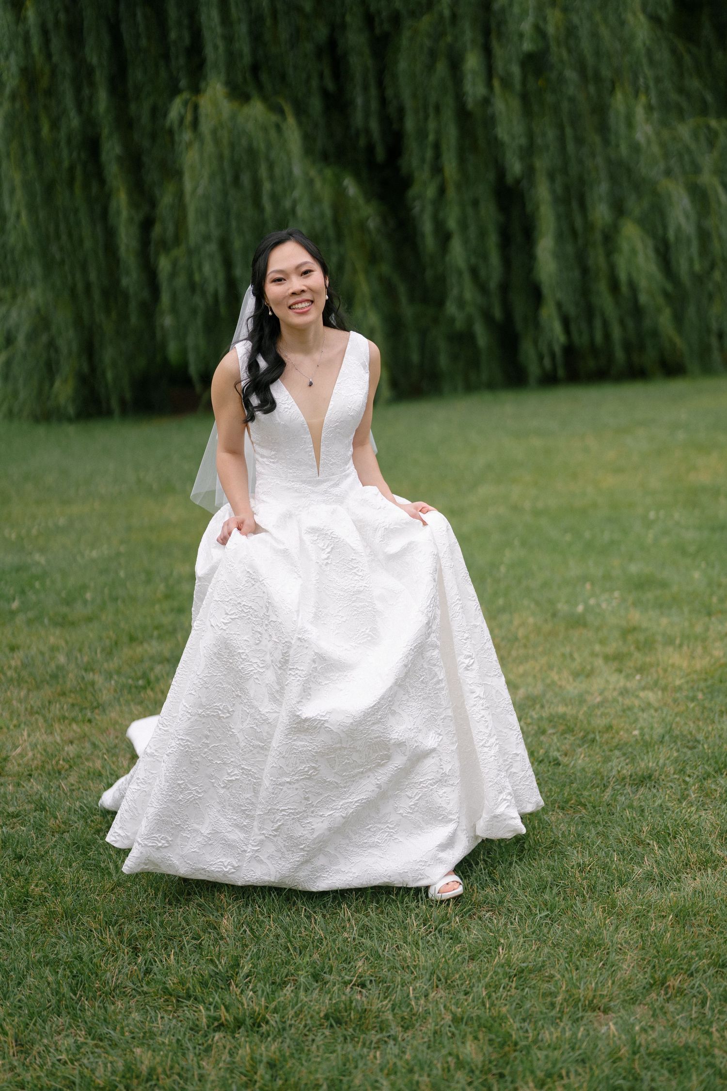 Smiling Asian bride in deep V-neck white ballgown lifts skirt while posing near weeping willow tree.