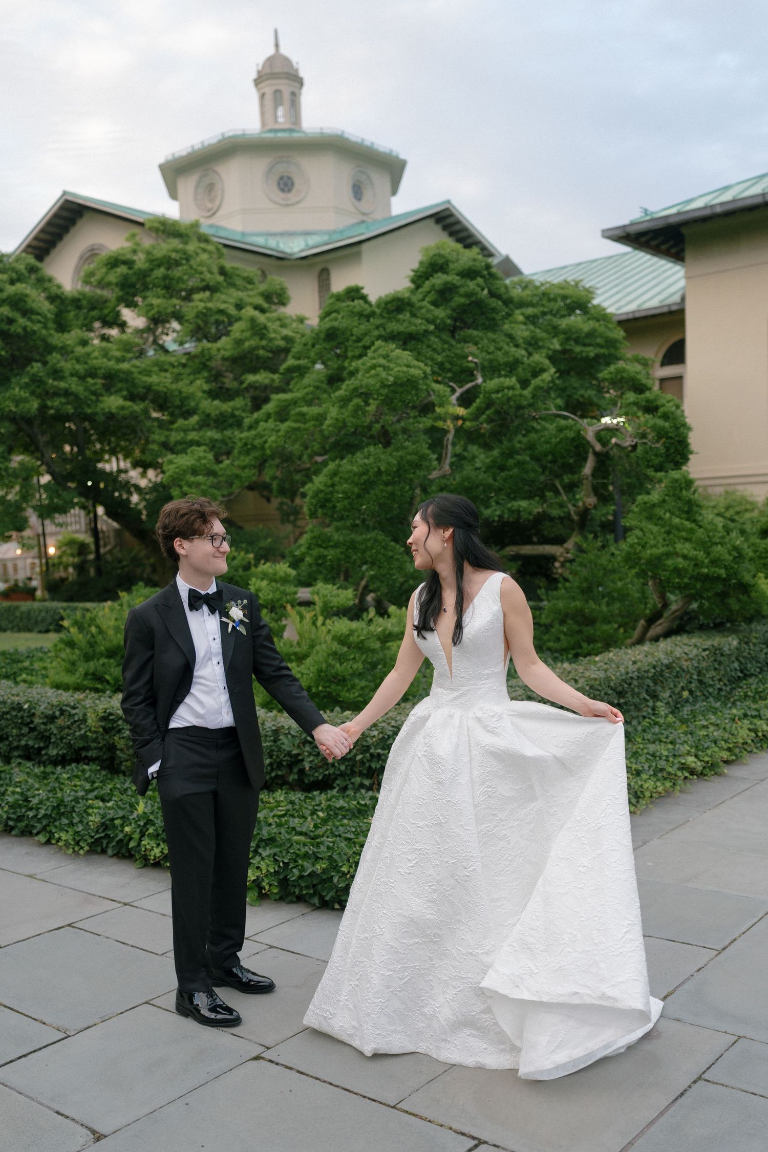 Bride and groom hold hands near domed building in garden.