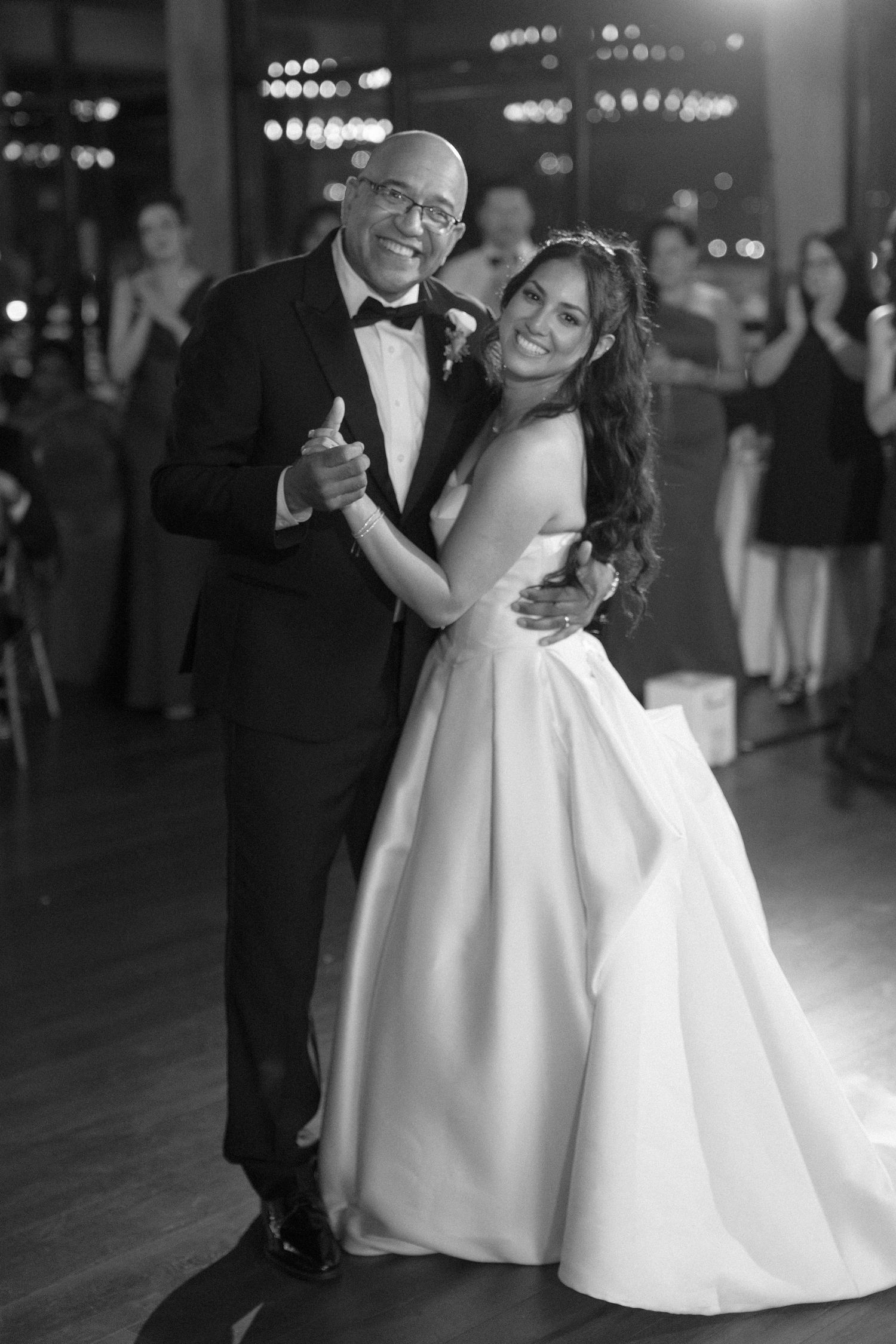 Black and white photo of a bride and groom sharing a joyful father-daughter dance at an elegant wedding reception.