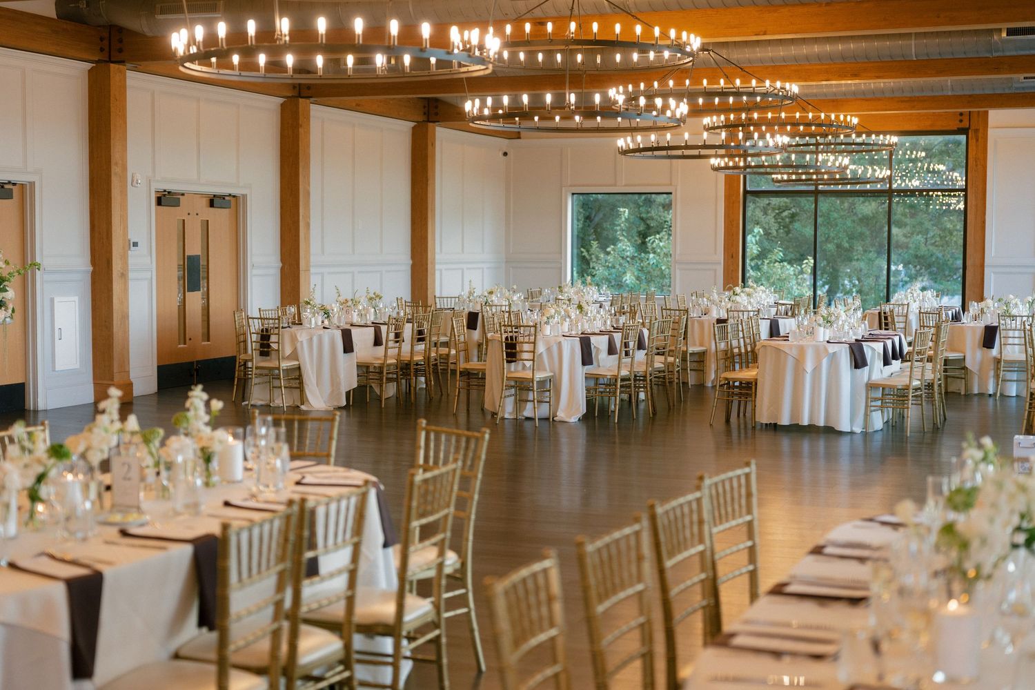 Elegant wedding reception hall with gold chiavari chairs, round tables with white linens, and warm chandelier lighting.