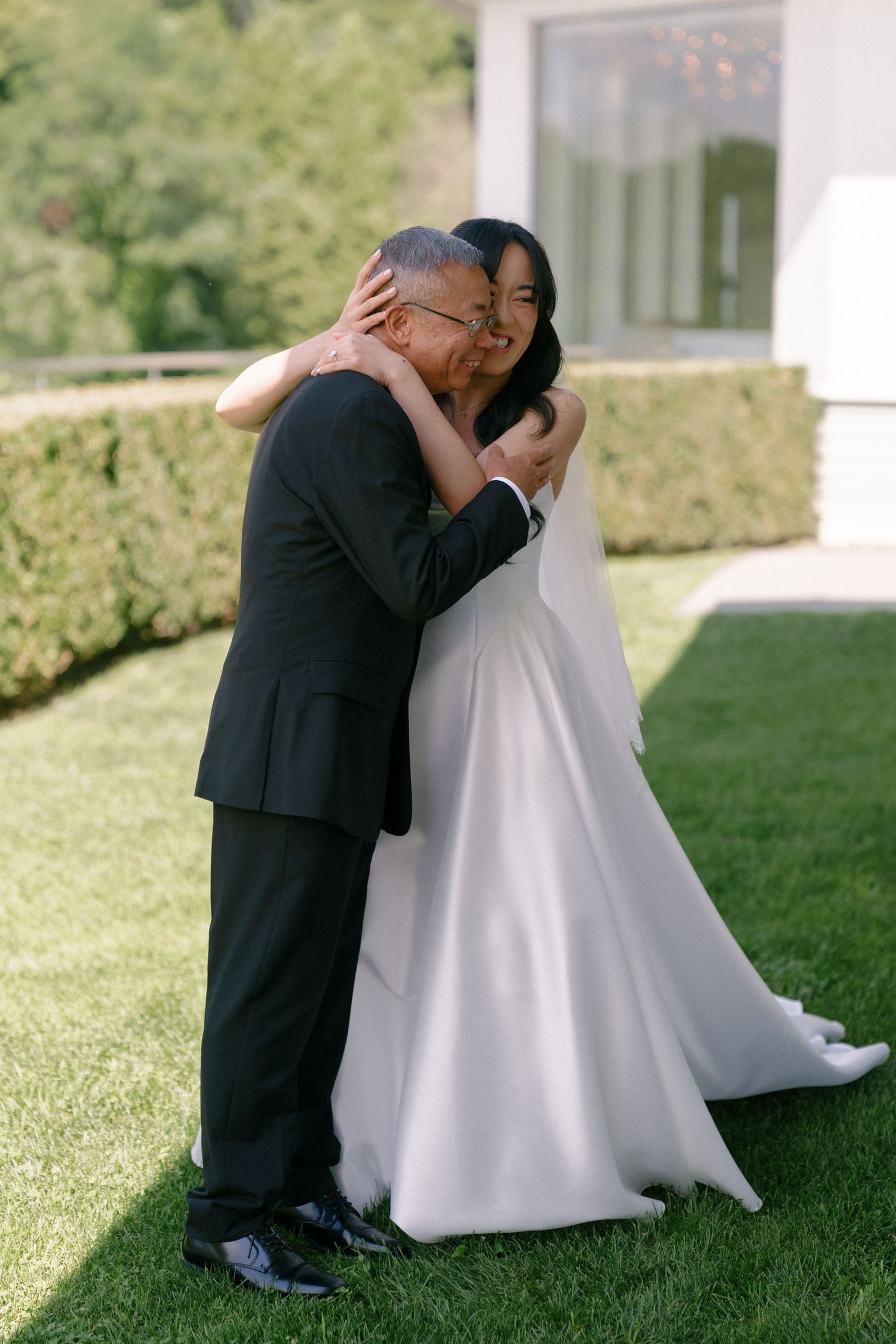 Emotional father-daughter first look moment with bride in white gown embracing on green lawn.