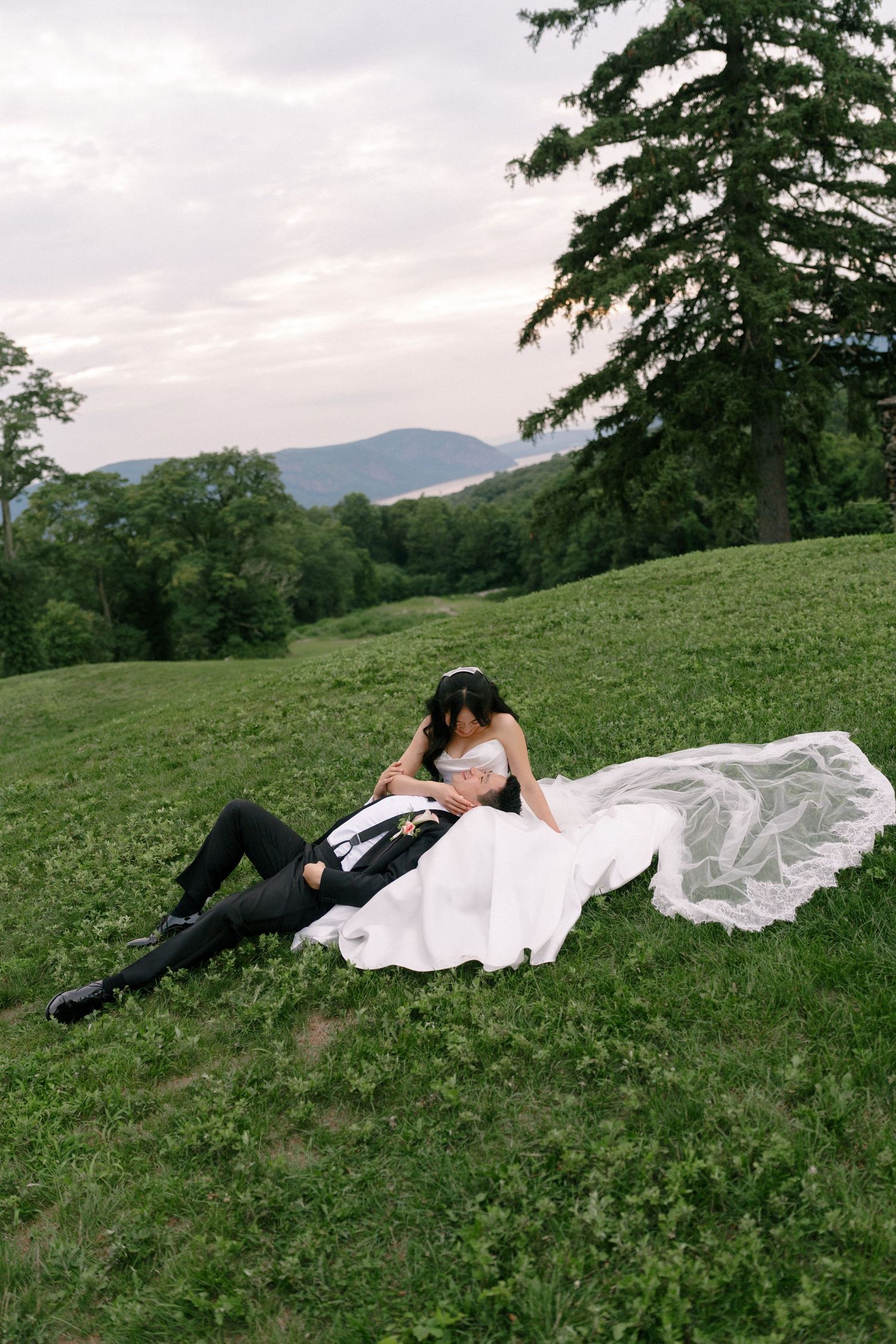 Bride and groom relax on a lush green hillside with mountains in the background, bride's veil flowing in the breeze.
