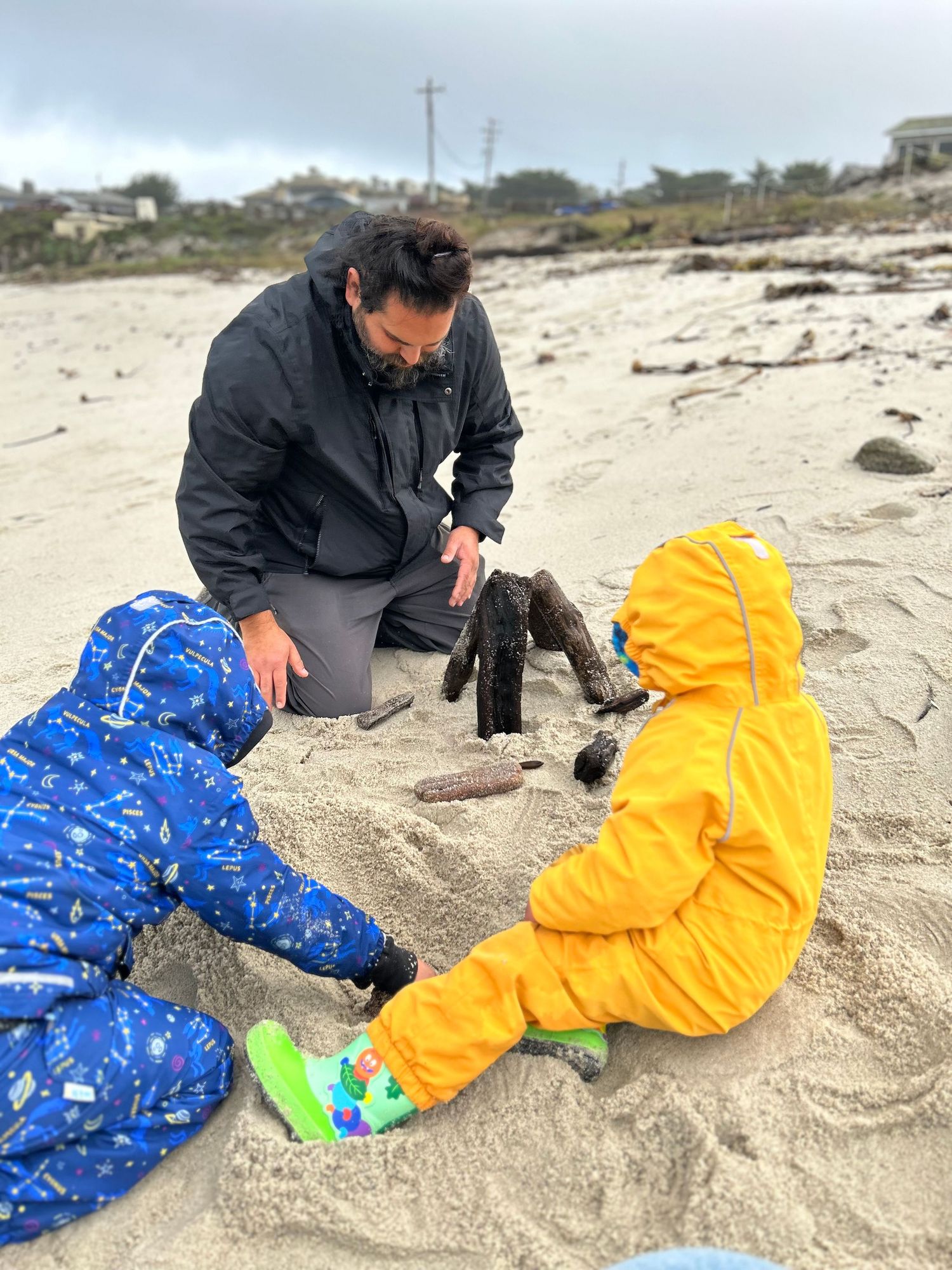 A family plays together on a sandy beach with children wearing colorful rain suits in blue and yellow.