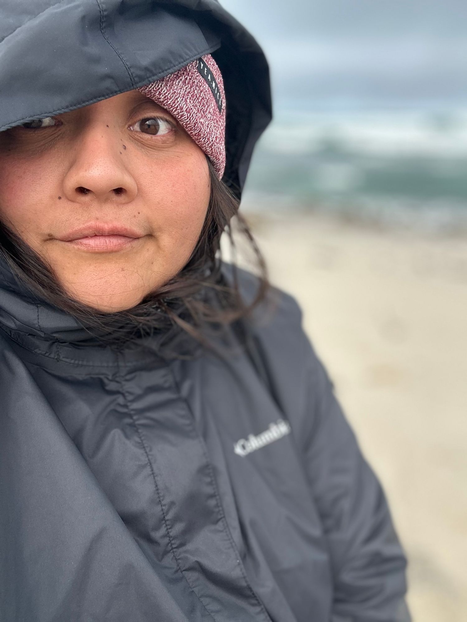 Person wearing a gray jacket and pink beanie looks to the side while at a beach on an overcast day.