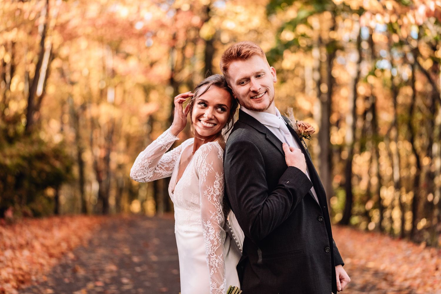 A couple poses together on a leaf-covered path in the woods during autumn with vibrant orange foliage in background.