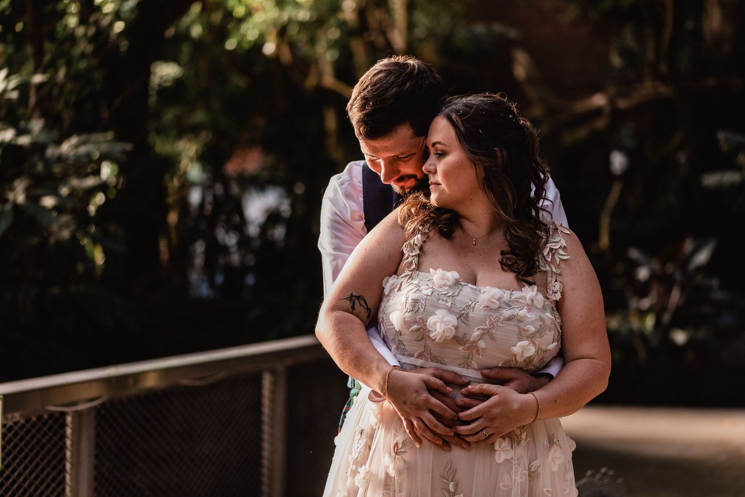 A romantic couple shares an intimate moment on a wooden bridge with sunlit foliage in the background.
