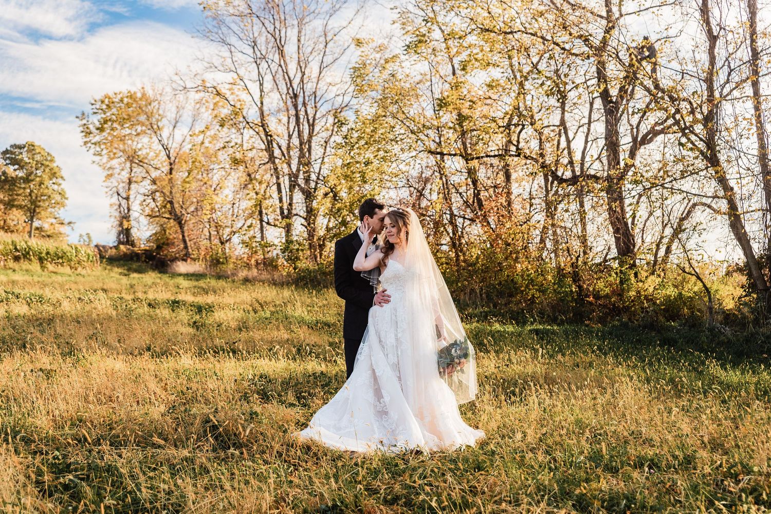 A couple shares an intimate moment in a sunlit autumn meadow with golden trees and grass in the background.