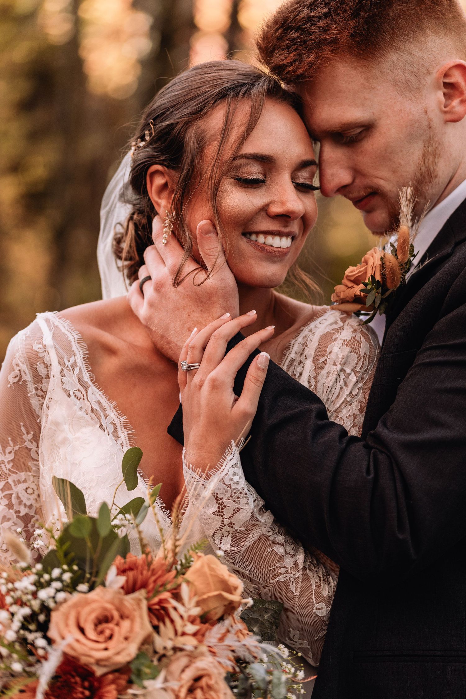 A romantic wedding photo of a couple embracing outdoors with autumn colors and a peach rose bouquet.