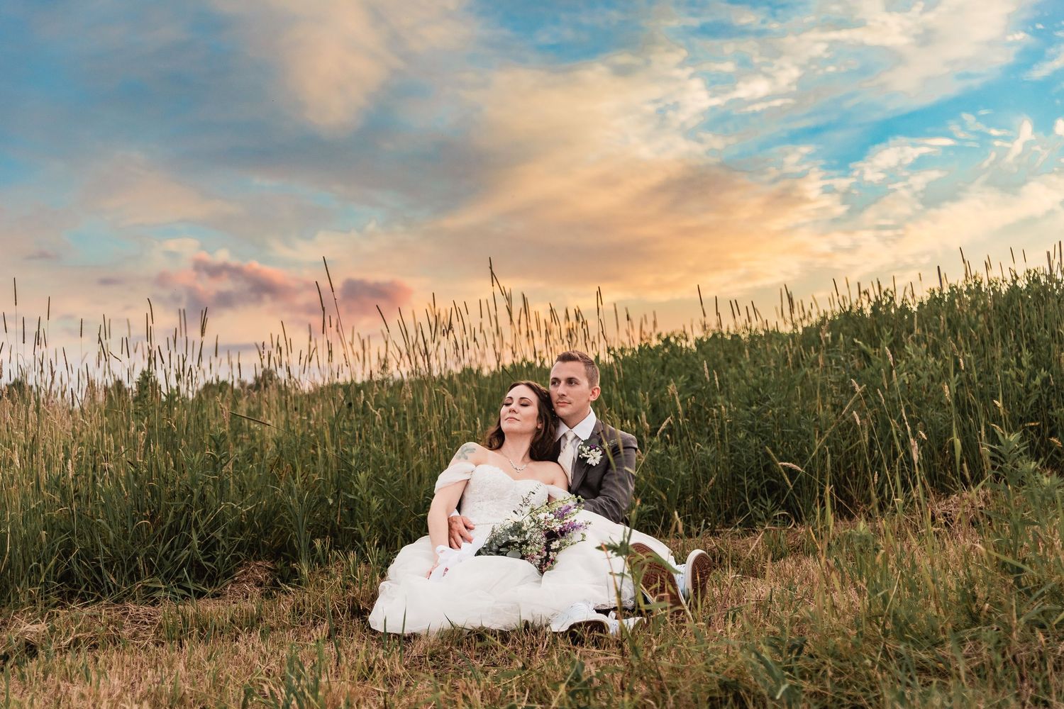 A couple in wedding attire sits together in a grassy field during sunset with dramatic orange and blue sky overhead.