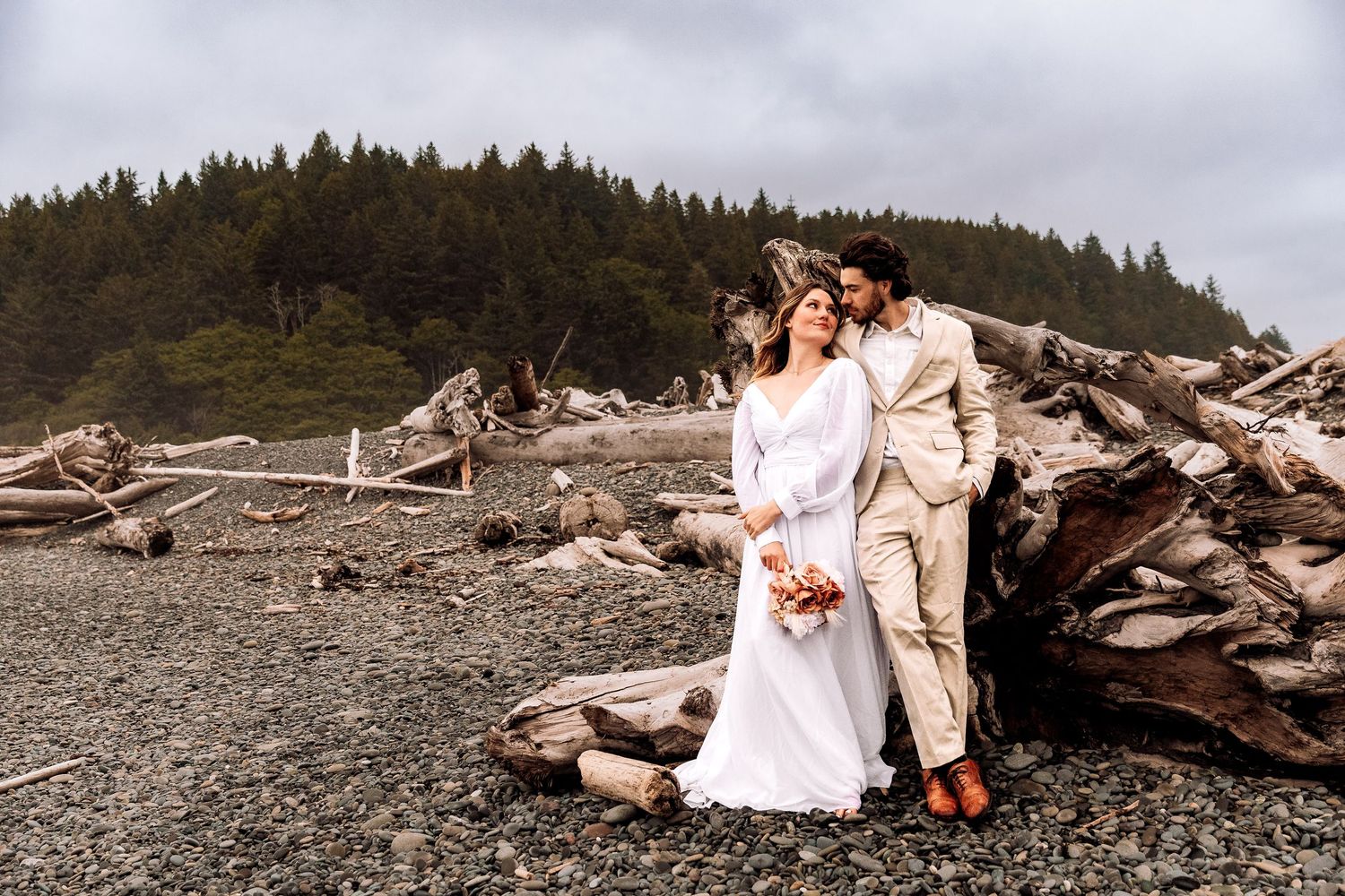 A wedding photoshoot on a rocky beach with driftwood and evergreen forest backdrop on an overcast day.