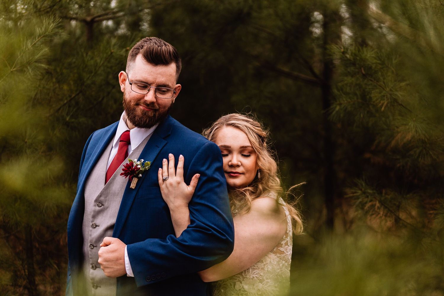 A bride and groom share a romantic moment in a wooded outdoor setting, wearing navy blue and white wedding attire.