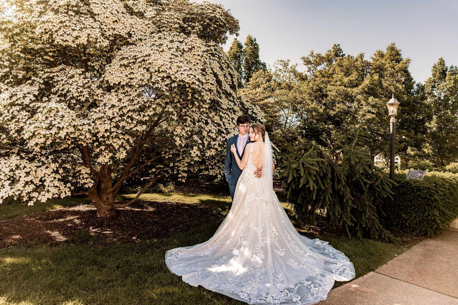 A couple embraces under a flowering tree in a garden setting, with the bride wearing a flowing lace gown with long train.