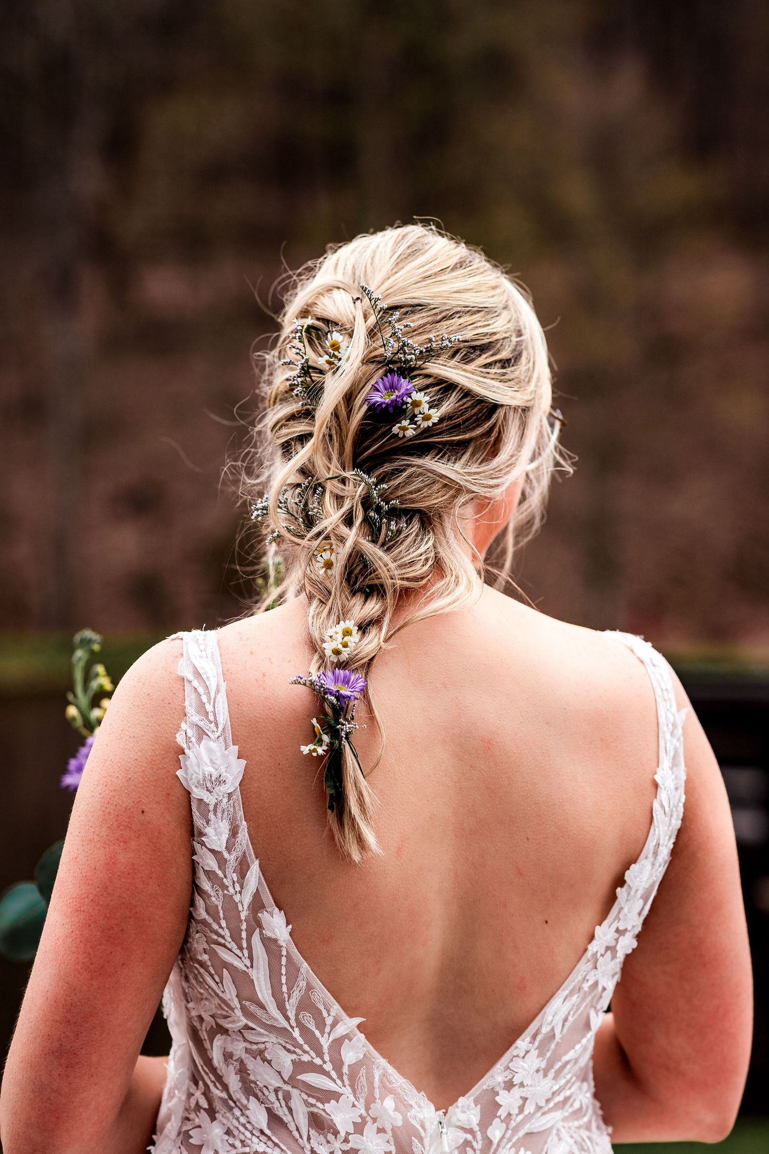 Elegant bridal hairstyle with purple flowers woven into a loose braid, shown from behind with lace wedding dress detail.