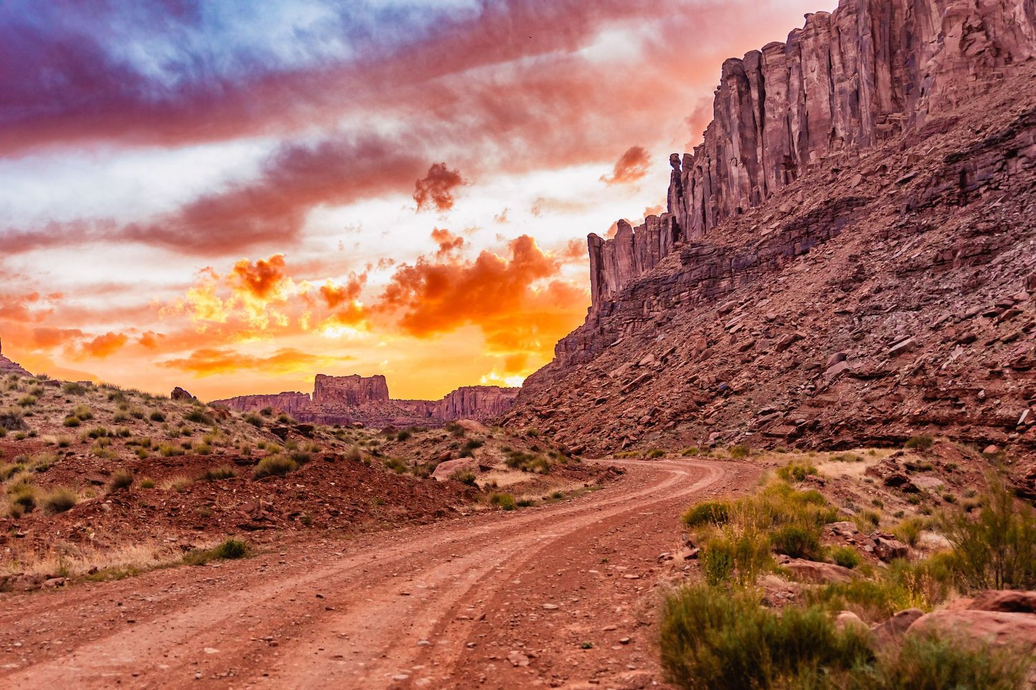 A winding dirt road leads through red rock cliffs in Monument Valley during a dramatic orange desert sunset.