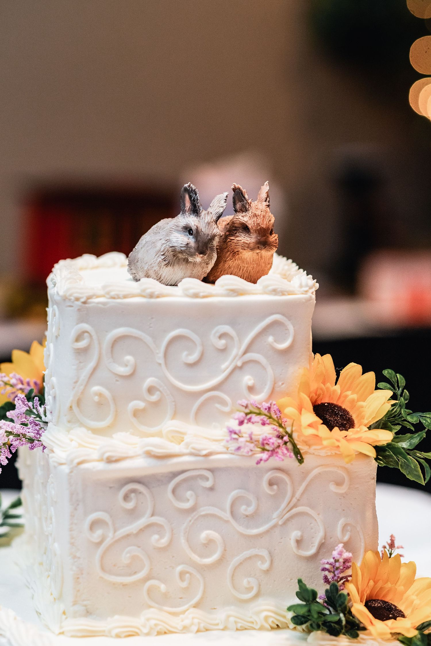 Two-tiered white wedding cake with scrollwork design decorated with sunflowers and topped with ceramic bunny figurines.