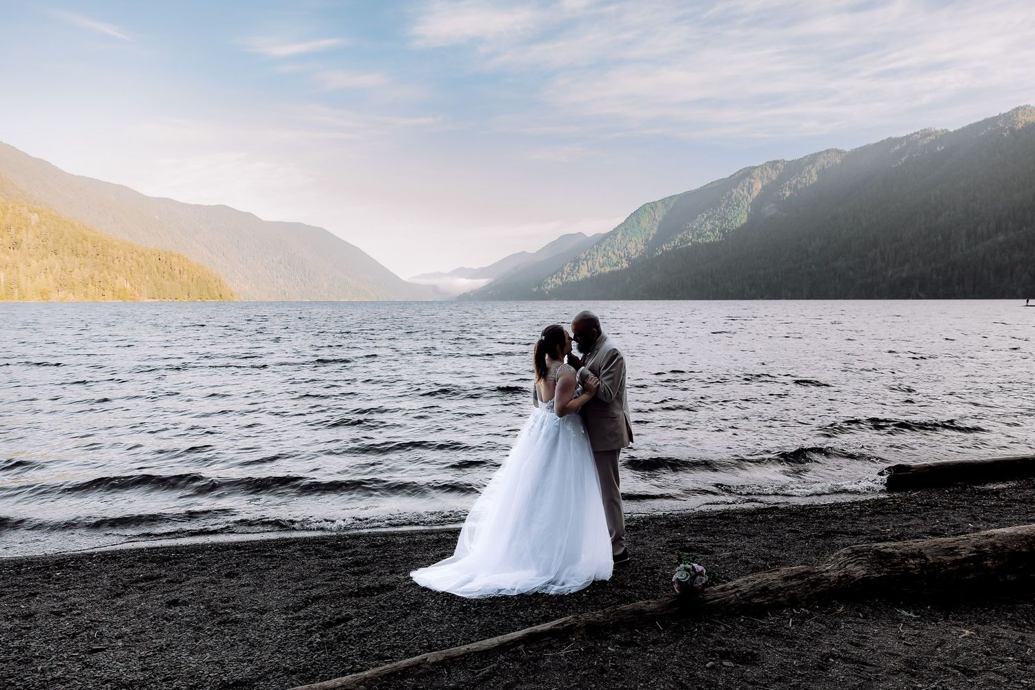Silhouetted couple in wedding attire embrace by a serene mountain lake at sunset with dramatic mountain backdrop.