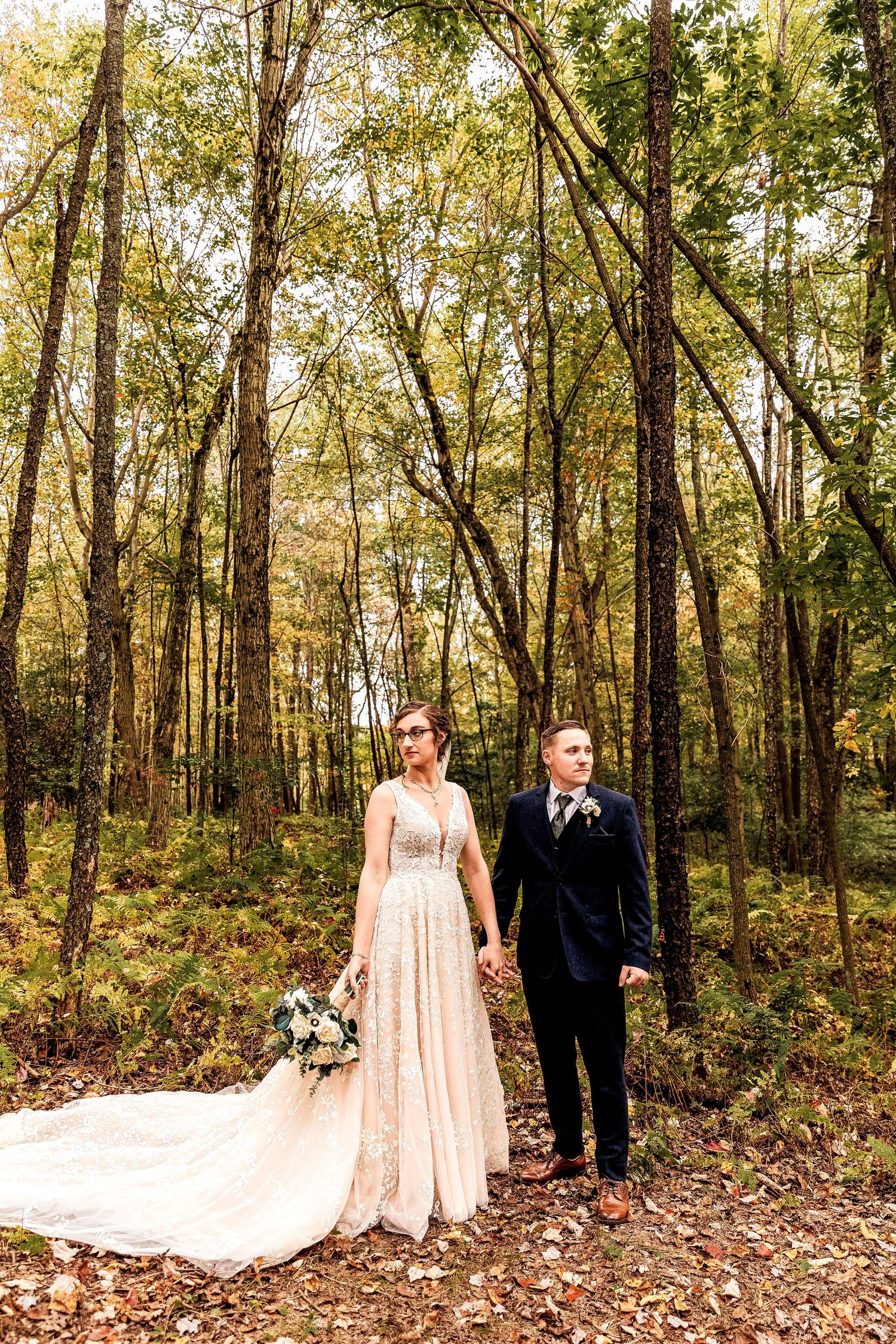 A romantic outdoor wedding photo showing a couple in formal attire standing together on a leaf-covered forest path.