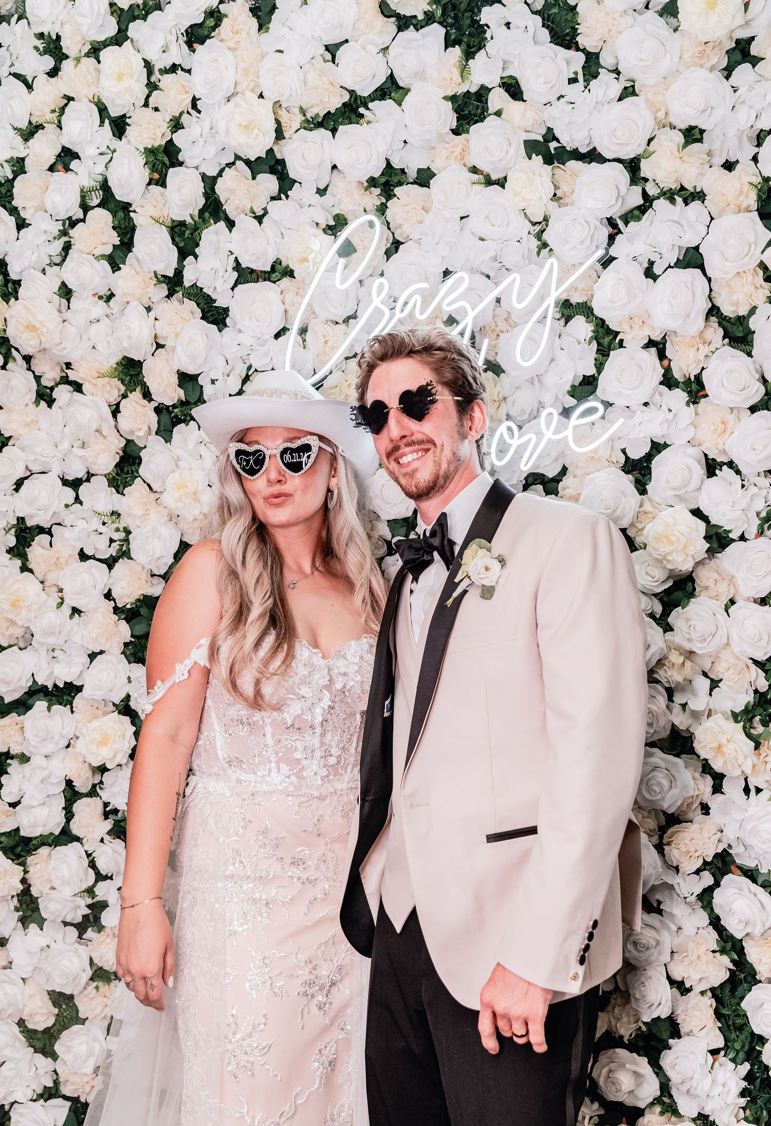 Bride and groom at small intimate wedding pose in front of white rose flower wall with neon “crazy love” sign, wearing fun sunglasses