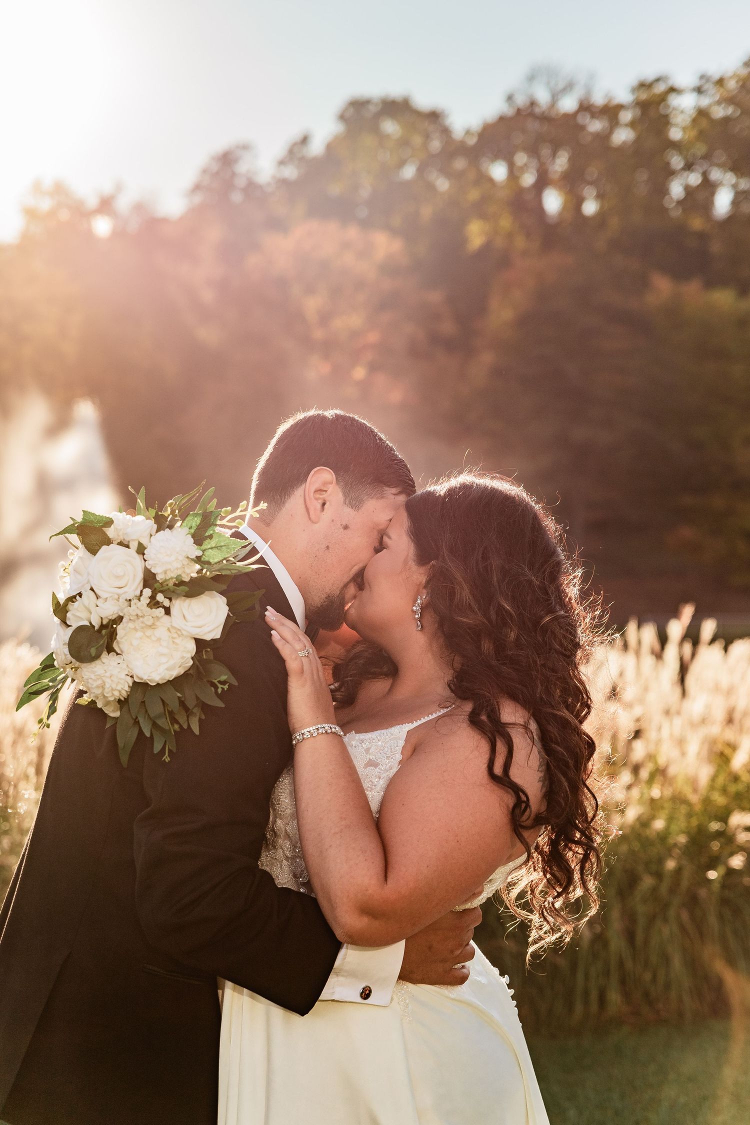 Newlywed couple shares romantic kisses in front of golden sunlit field of pampas grass at sunset.