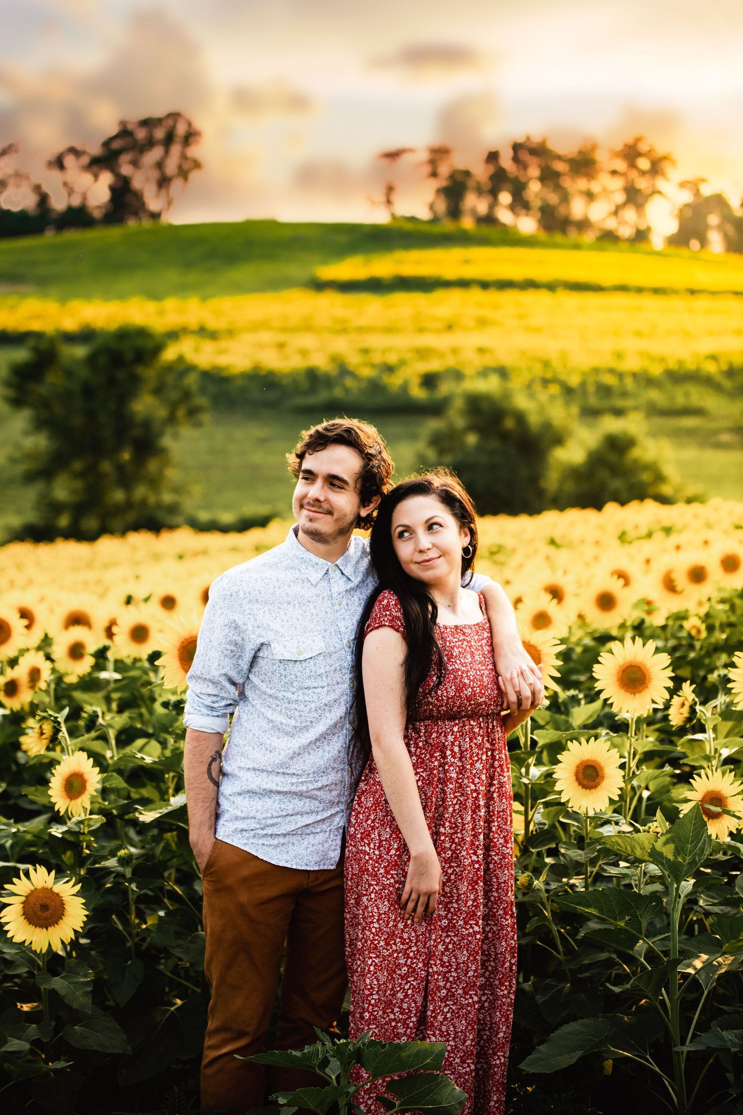 A romantic couple stands together in a vibrant sunflower field at golden hour with warm sunset lighting.