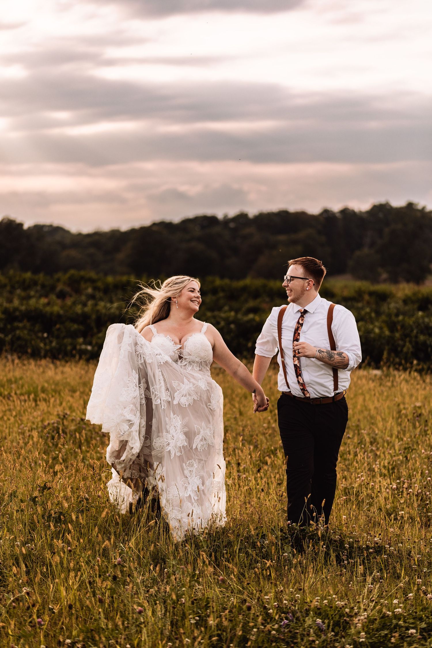 A couple walks hand in hand through a golden field at sunset, the bride in lace and groom in suspenders.