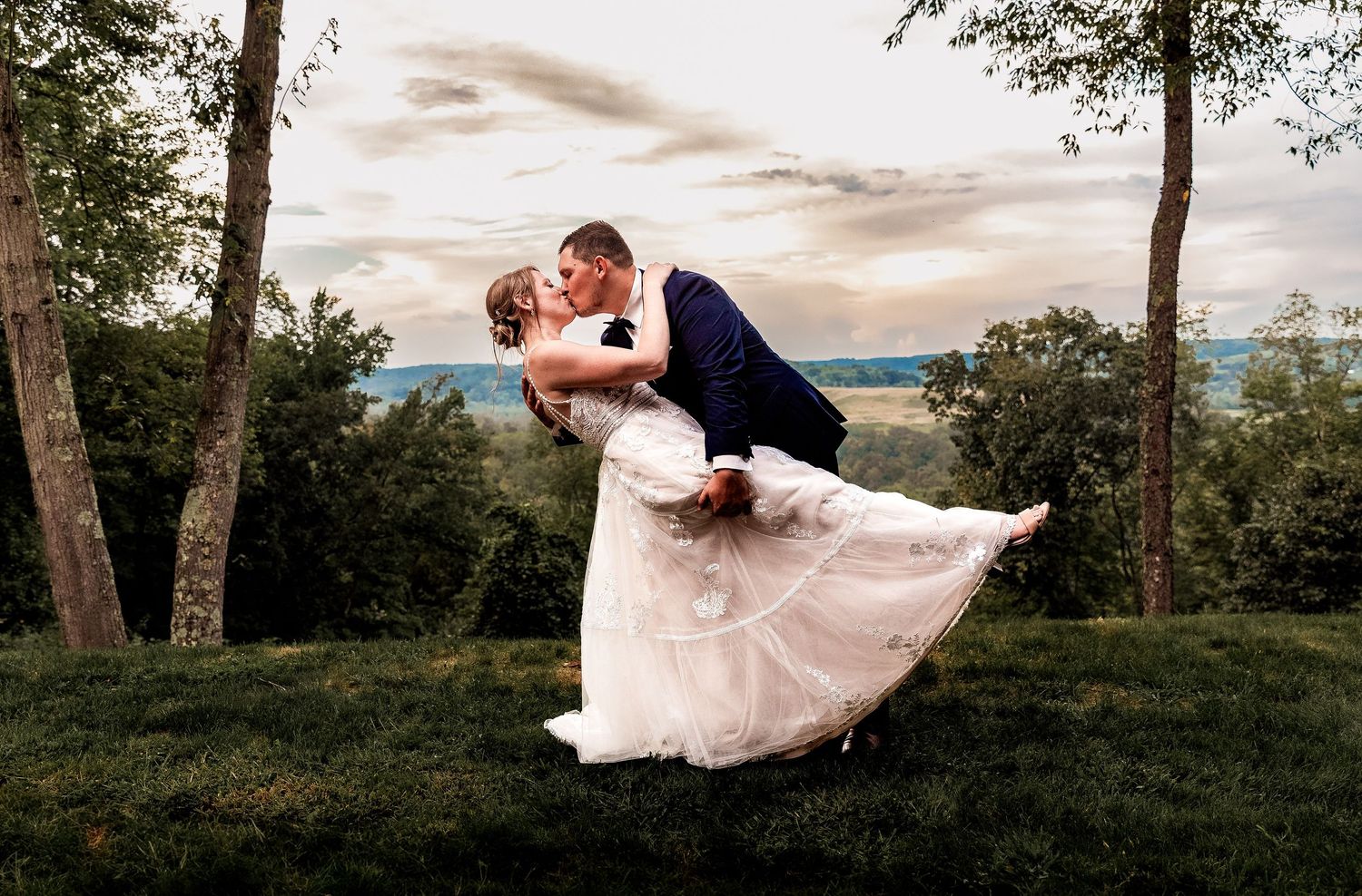 A romantic wedding couple embraces in a dramatic dip pose against a sunset mountain landscape.