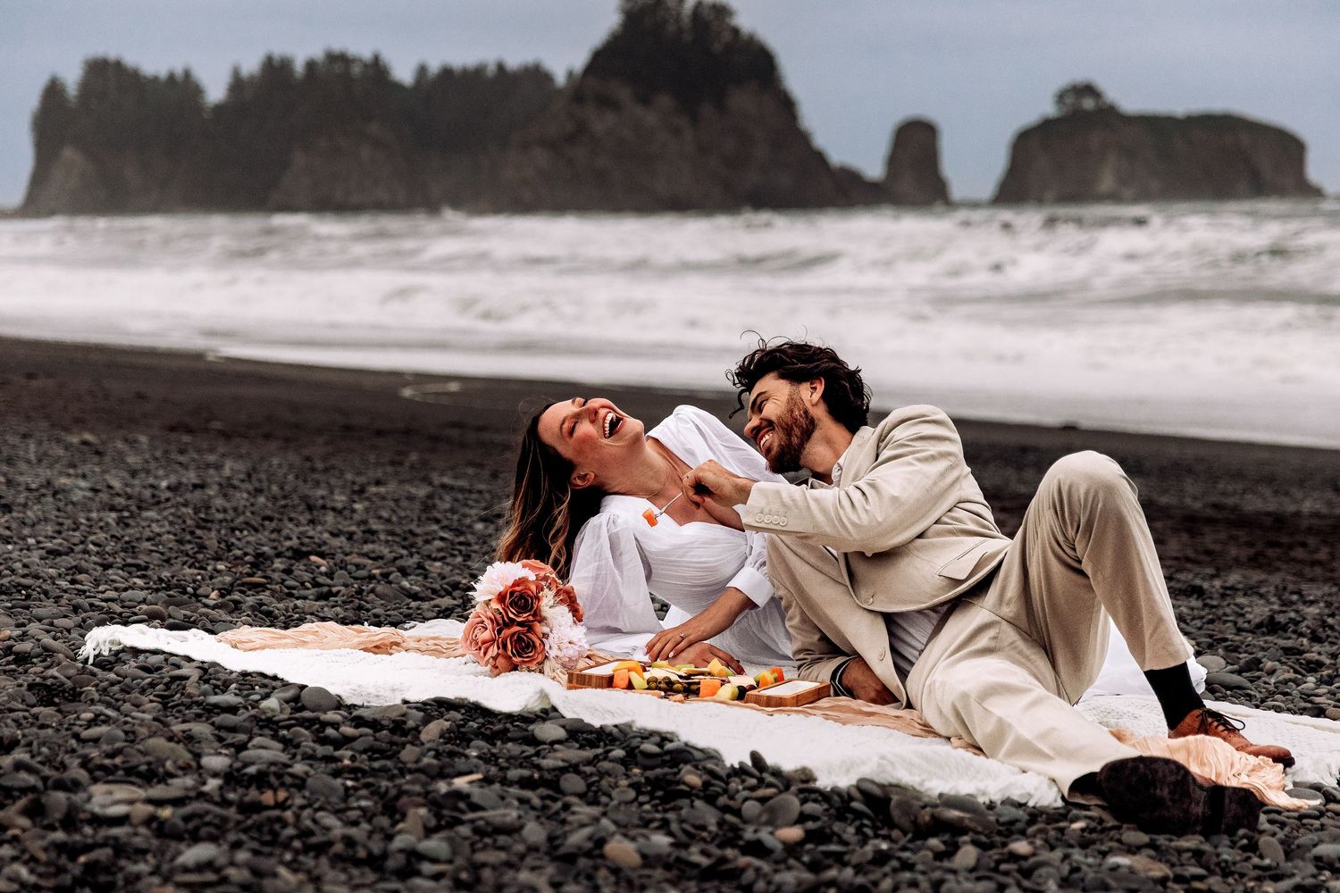Romantic couple enjoying picnic on black sand beach with dramatic coastal rock formations in background.