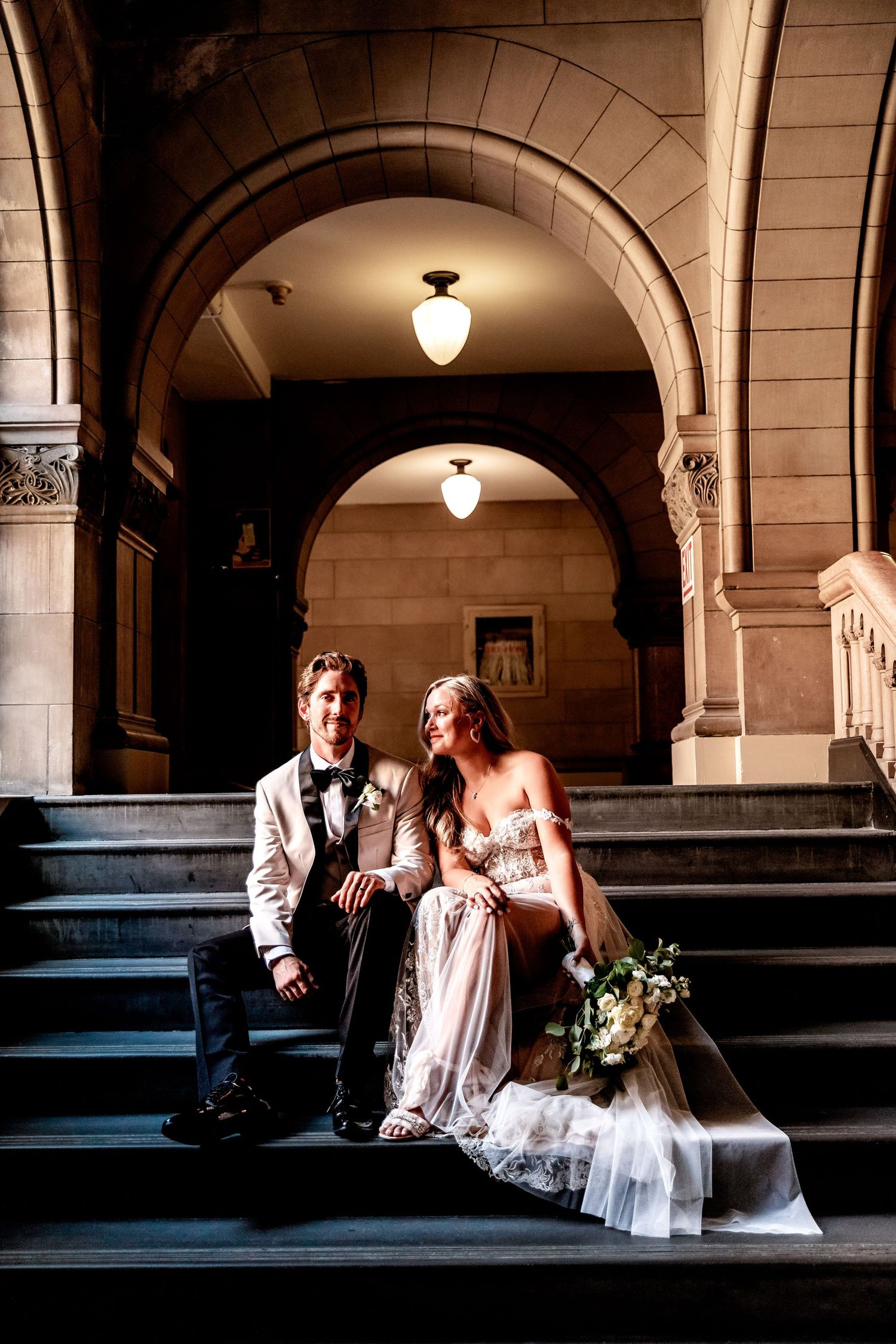 A romantic wedding couple sits under elegant stone archways in a classic architectural setting at dusk.