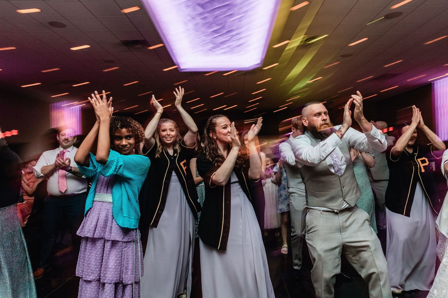 Wedding reception guests dancing together with hands raised under purple lighting at an indoor venue celebration.