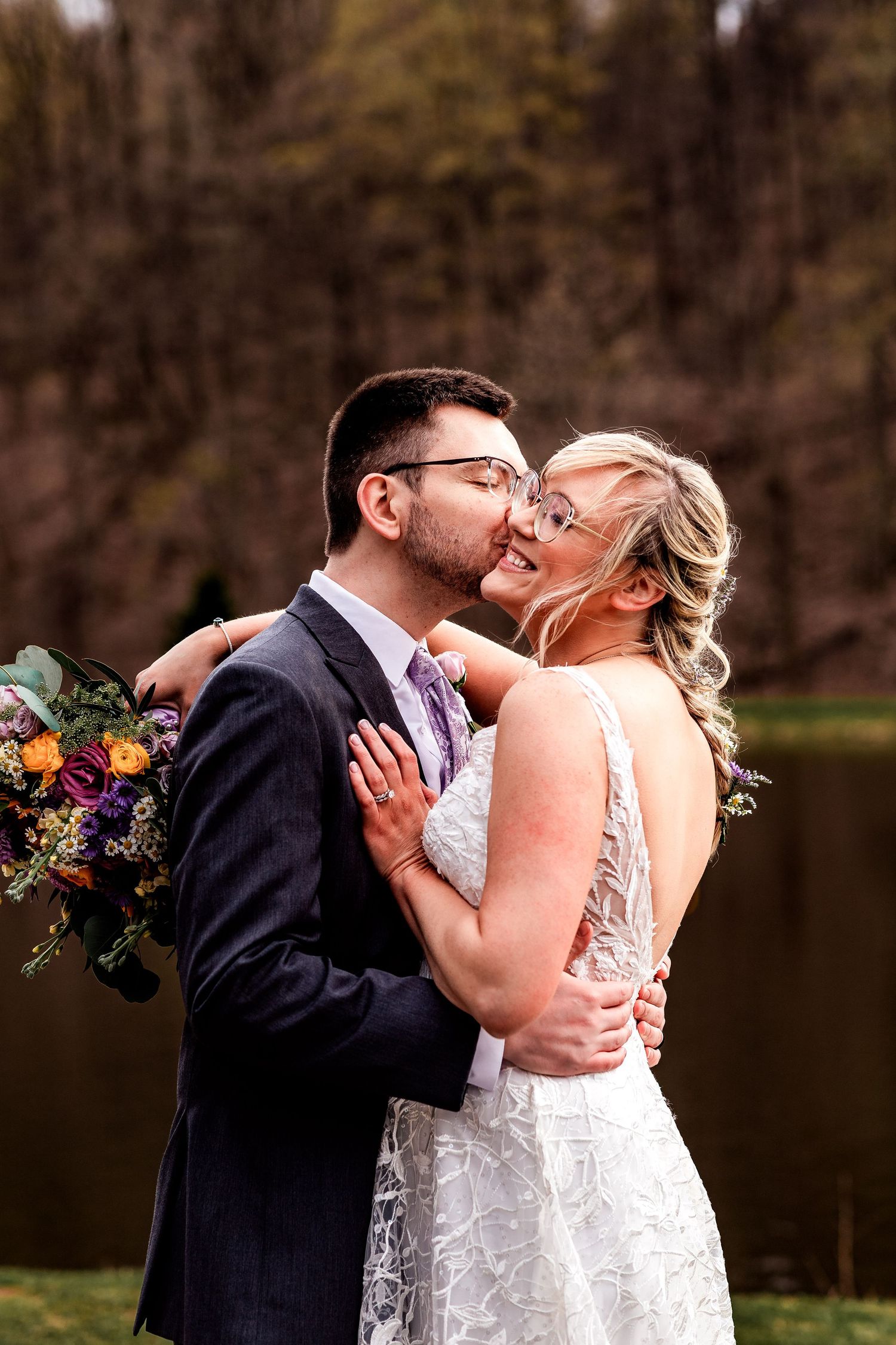 A bride in white lace holds a colorful bouquet while embracing and kissing outdoors with lake and trees in background.