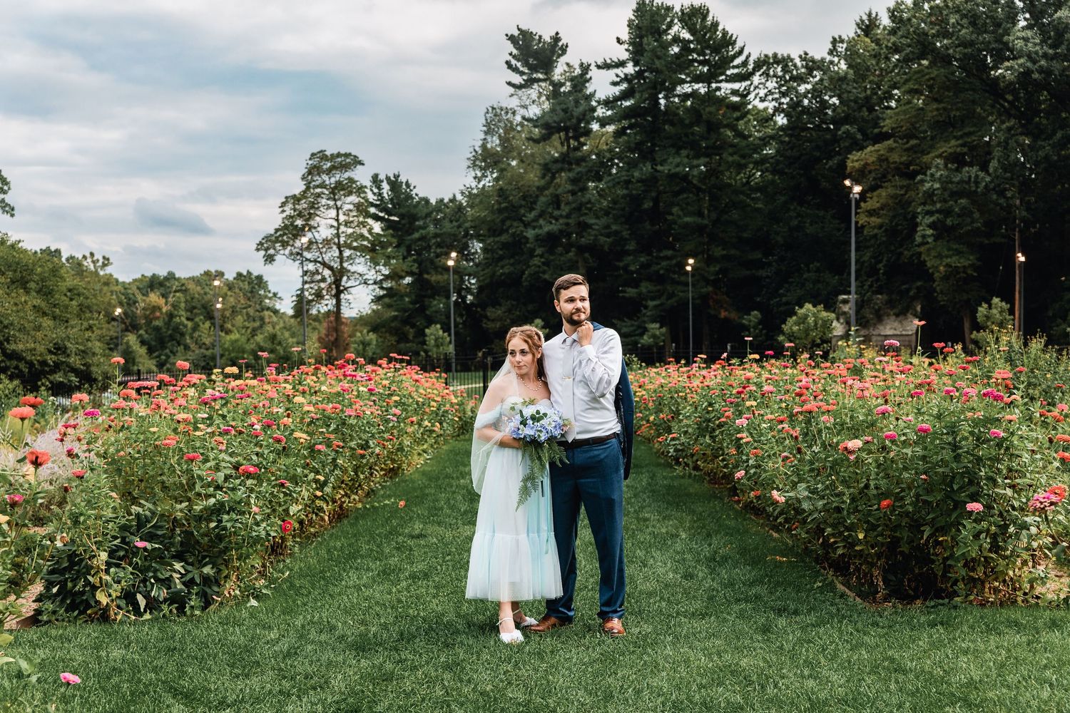 A couple stands on a green lawn path between vibrant flower gardens on a summer day.